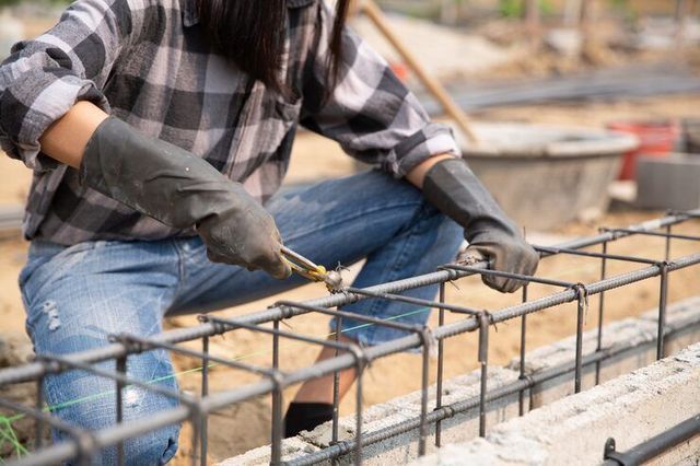 Woman in work clothes, wearing gloves, bending steel rebar at a construction site.