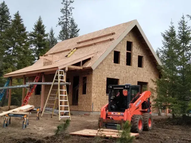 A house under construction with a partially built roof. An orange Kubota skid steer sits in front.
