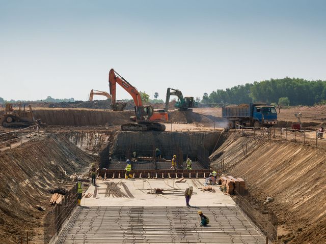 Construction site with excavators, workers, and a trench, likely for a new road or foundation. Blue sky and sunny day.