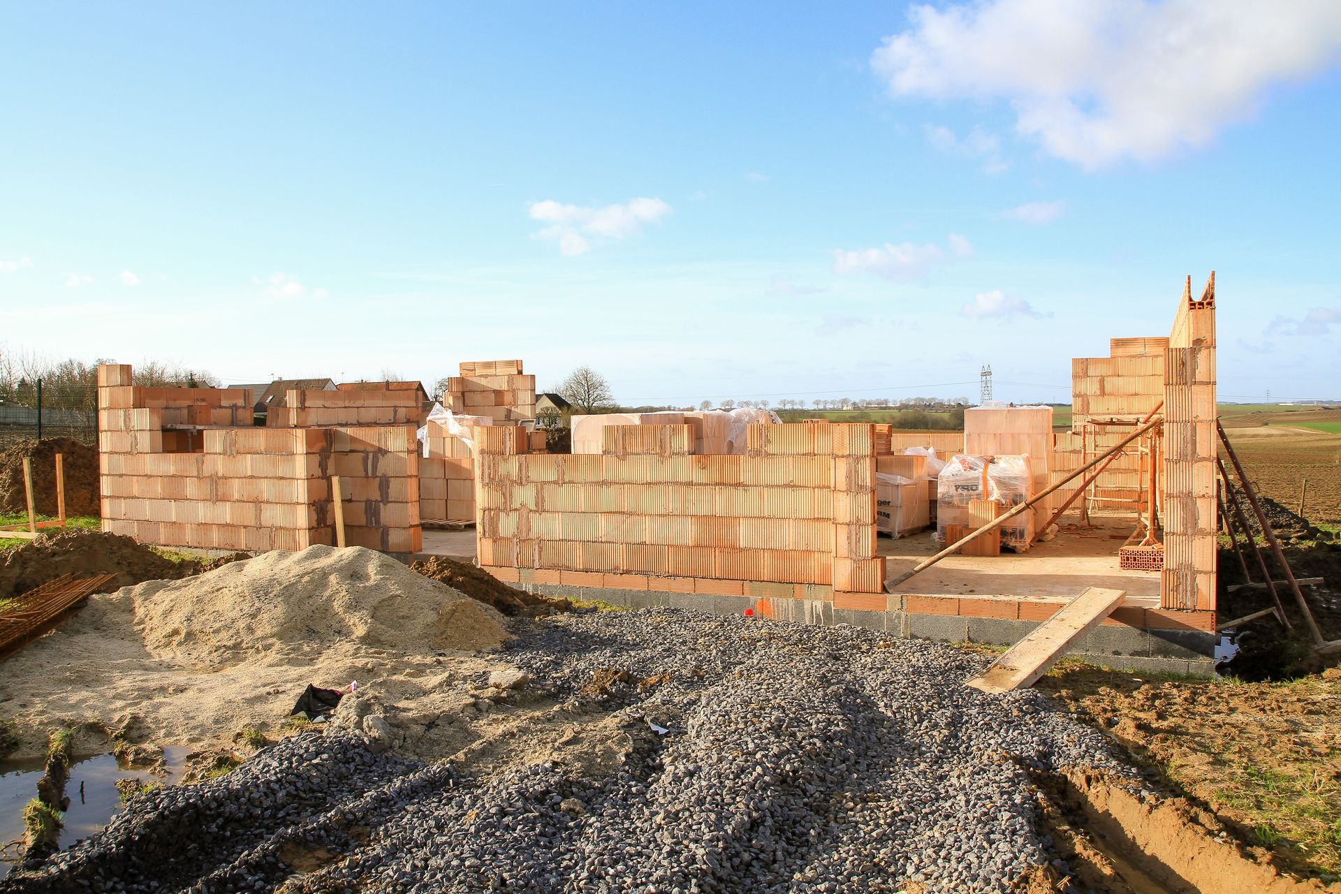Construction site with a brick building in progress, surrounded by dirt and construction materials under a blue sky.