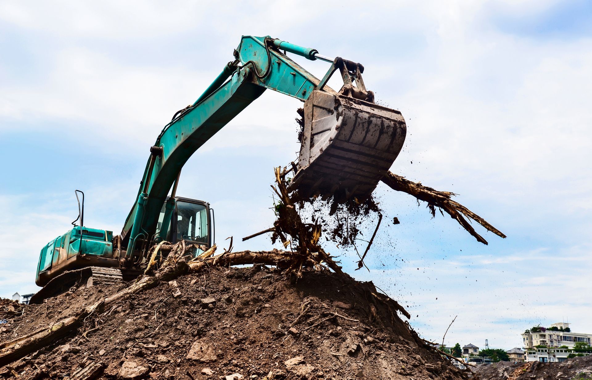 Green excavator removing tree roots from a dirt pile under a cloudy sky.