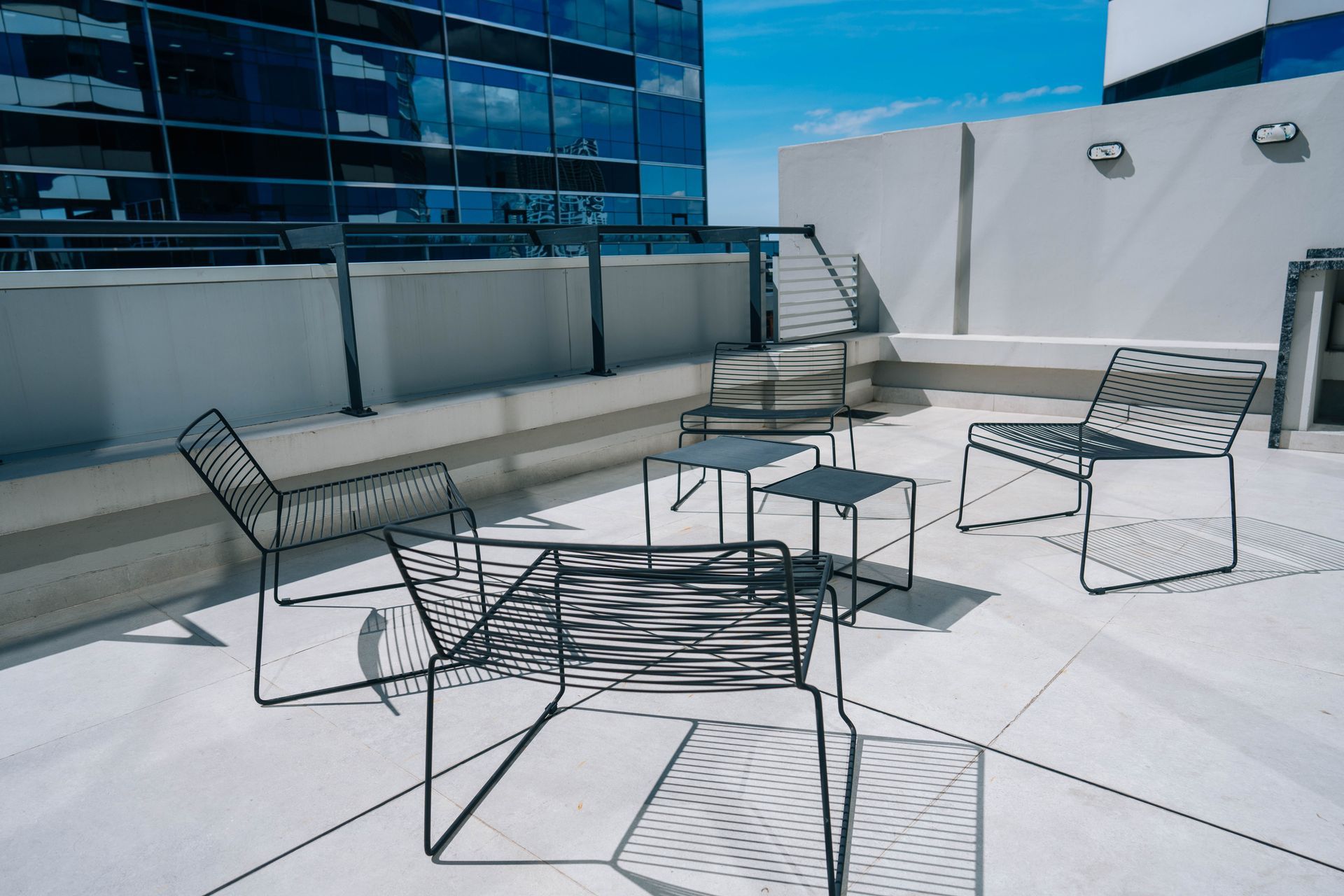 Patio en la azotea con sillas de metal negro y mesas pequeñas, con un edificio de cristal al fondo.