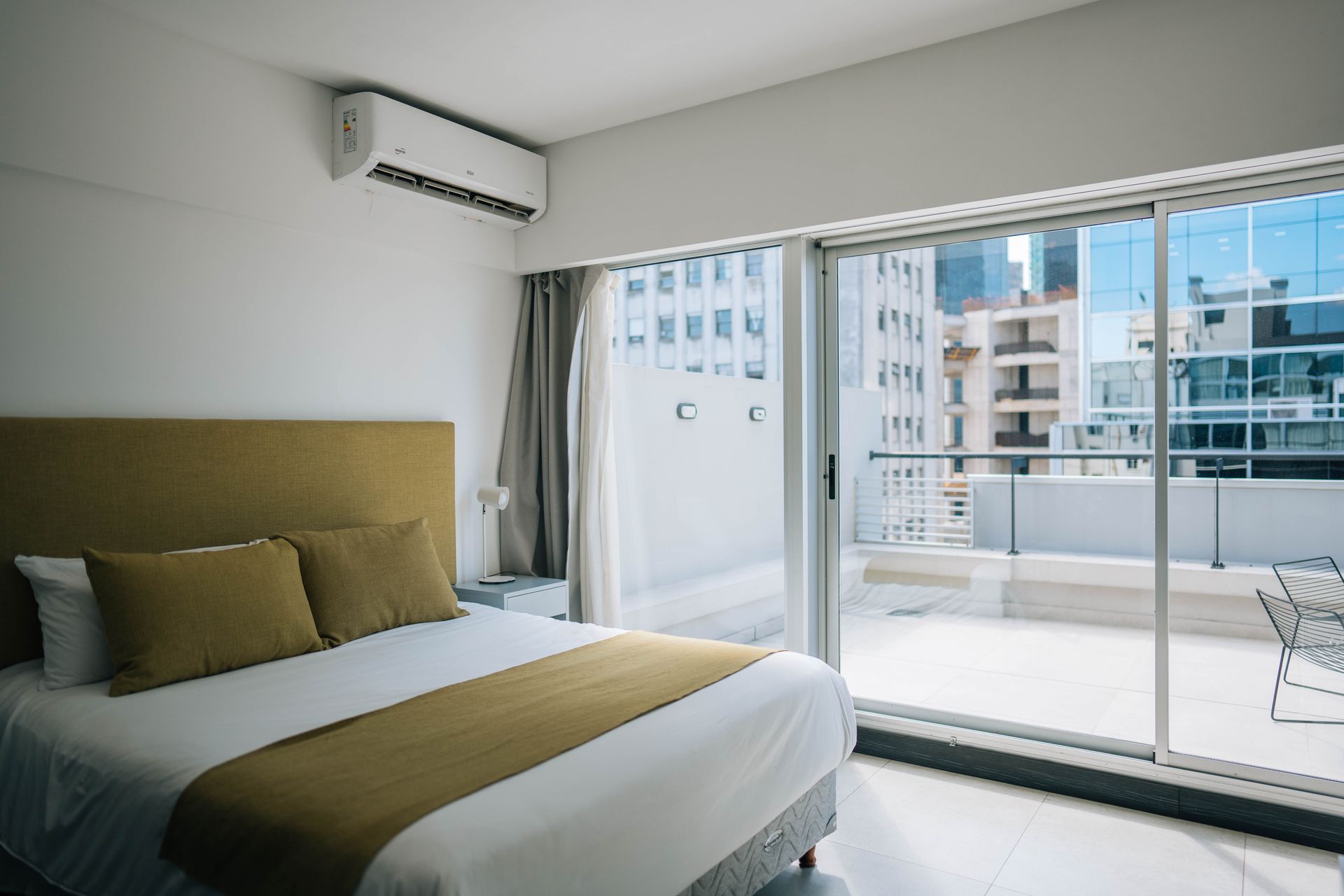 Dormitorio con cama, puerta corrediza de vidrio abierta al balcón, vista a la ciudad.