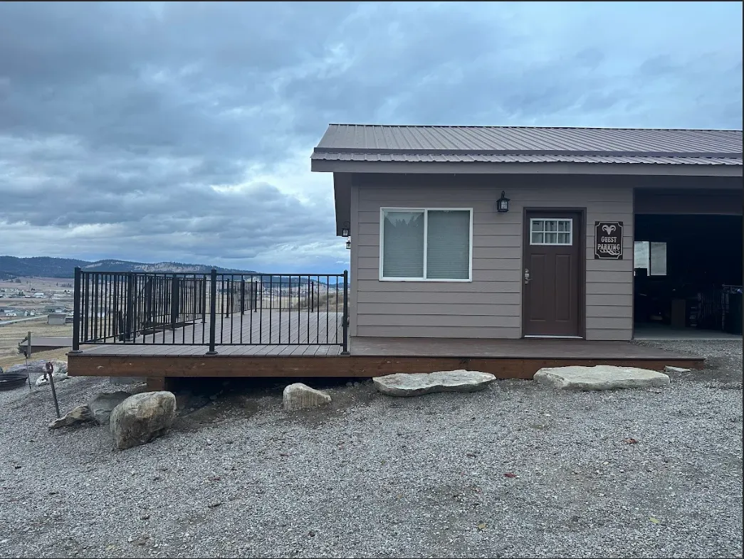 A tan house with a front deck and iron railing overlooks a distant valley under a cloudy sky.