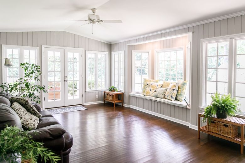 A bright sunroom with light gray paneled walls, dark wood floors, a dark sofa, a window seat with pillows, and potted plants.