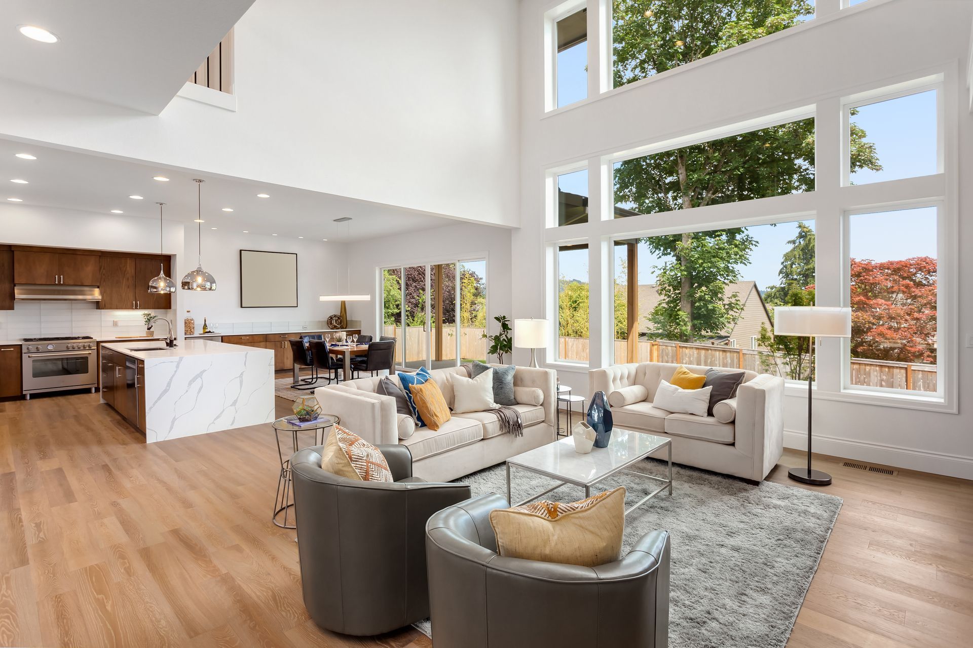 Open-concept living room with tall windows, a modern kitchen island, white sofas, and two grey armchairs on a rug.