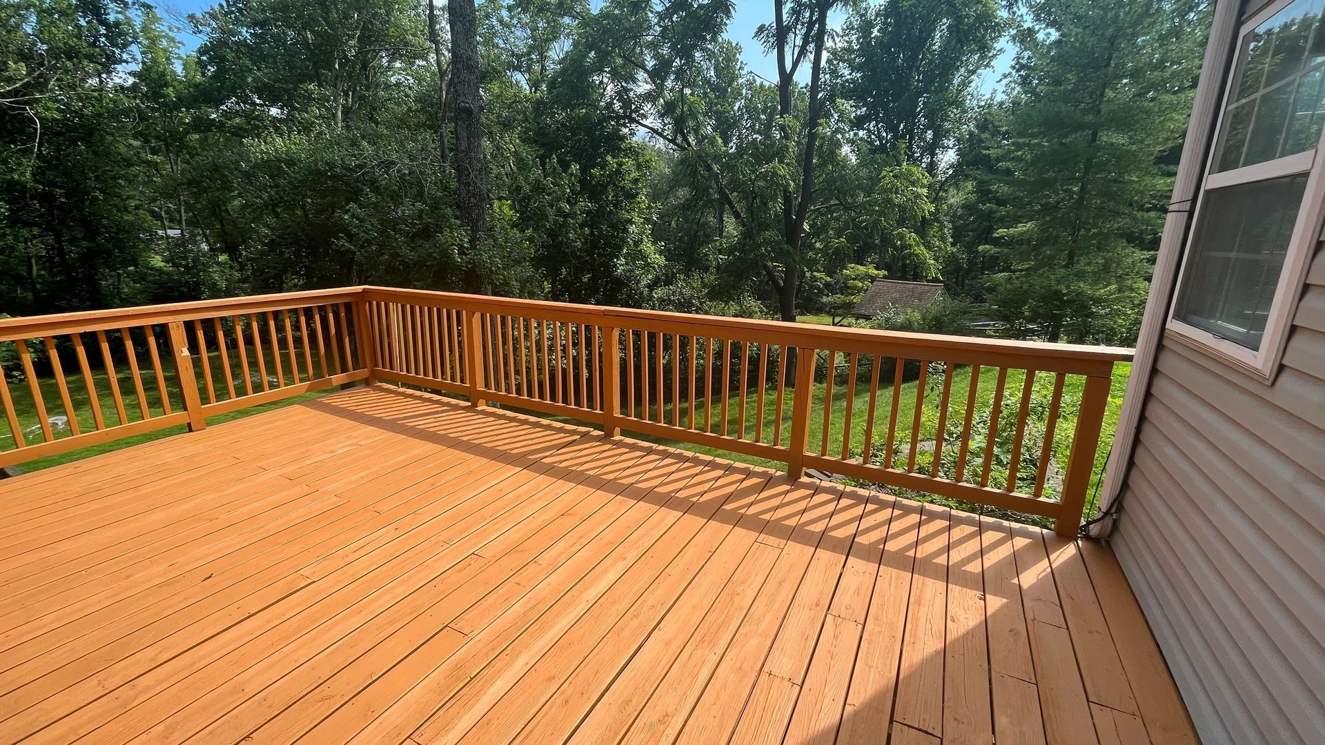 A large, freshly stained, orange-toned wooden deck with railings, overlooking a wooded yard on a sunny day.
