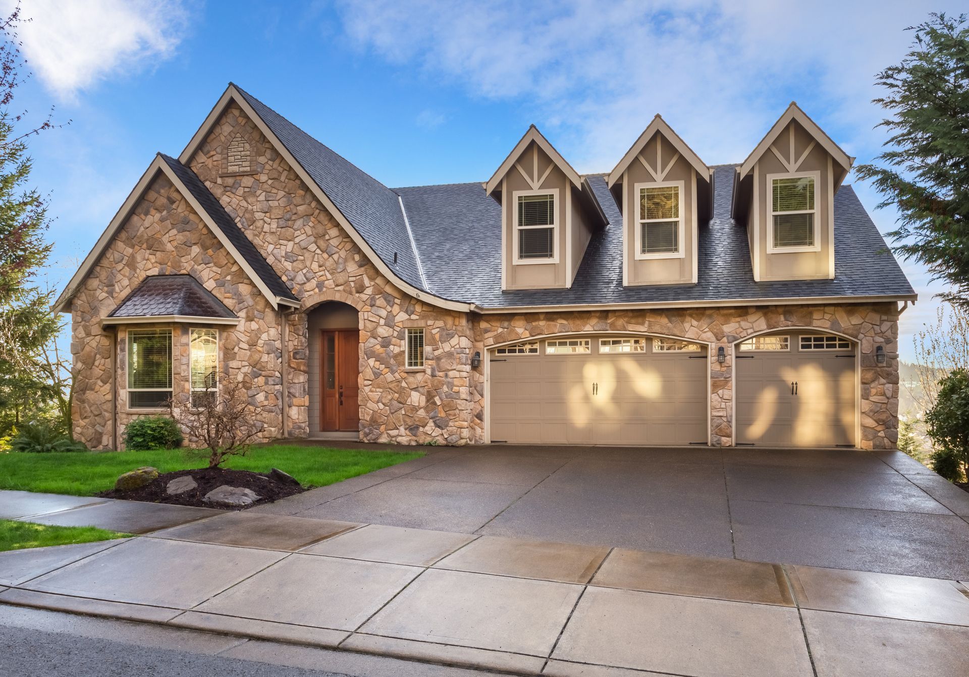 A stone-clad house with three dormer windows and a multi-car garage, fronted by a paved driveway and lawn.