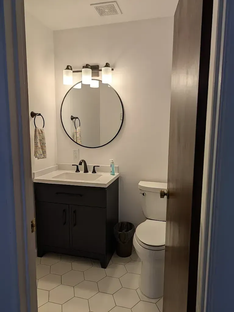 A small bathroom with a dark vanity, white hexagonal floor tiles, a round mirror, and a white toilet.