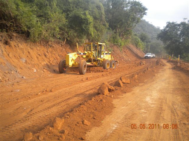 A yellow grader levels a dirt road. A car drives in the distance, with trees and hills in the background.