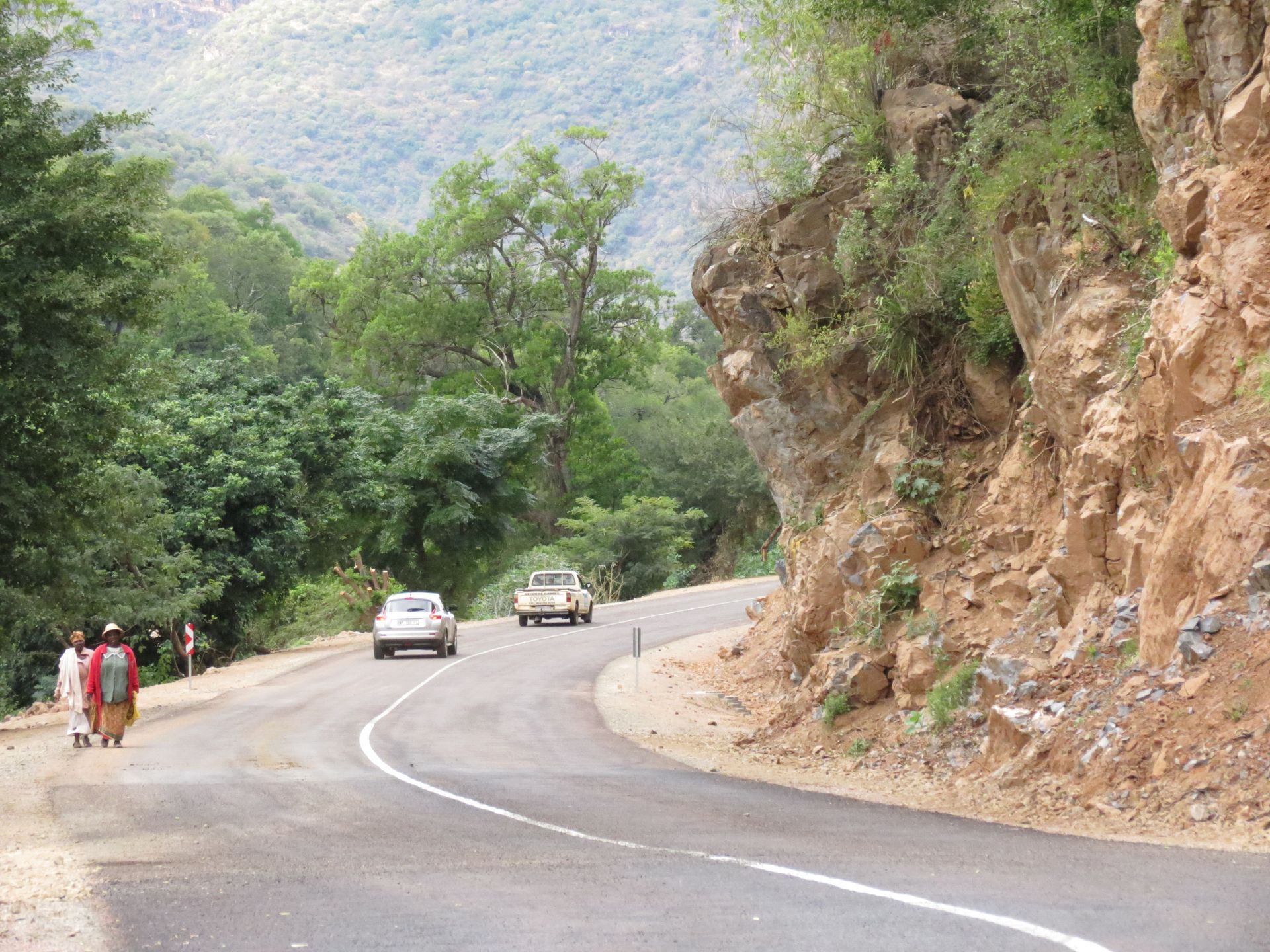 Winding road through a lush, green mountain landscape; cars and people walking on the side.