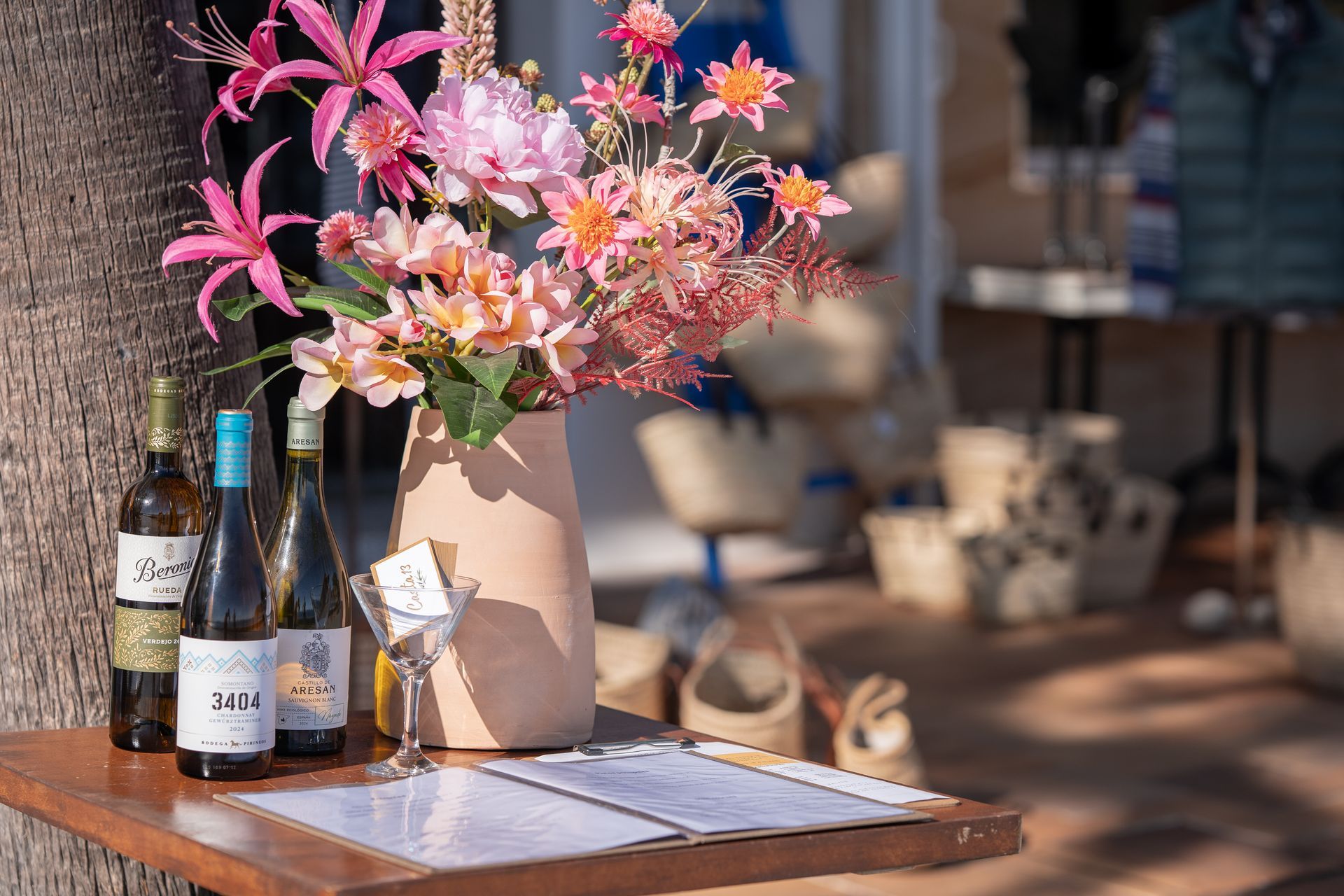 Mesa de madera con botellas de vino, flores, una copa de vino y un menú; entrada de la tienda al fondo.
