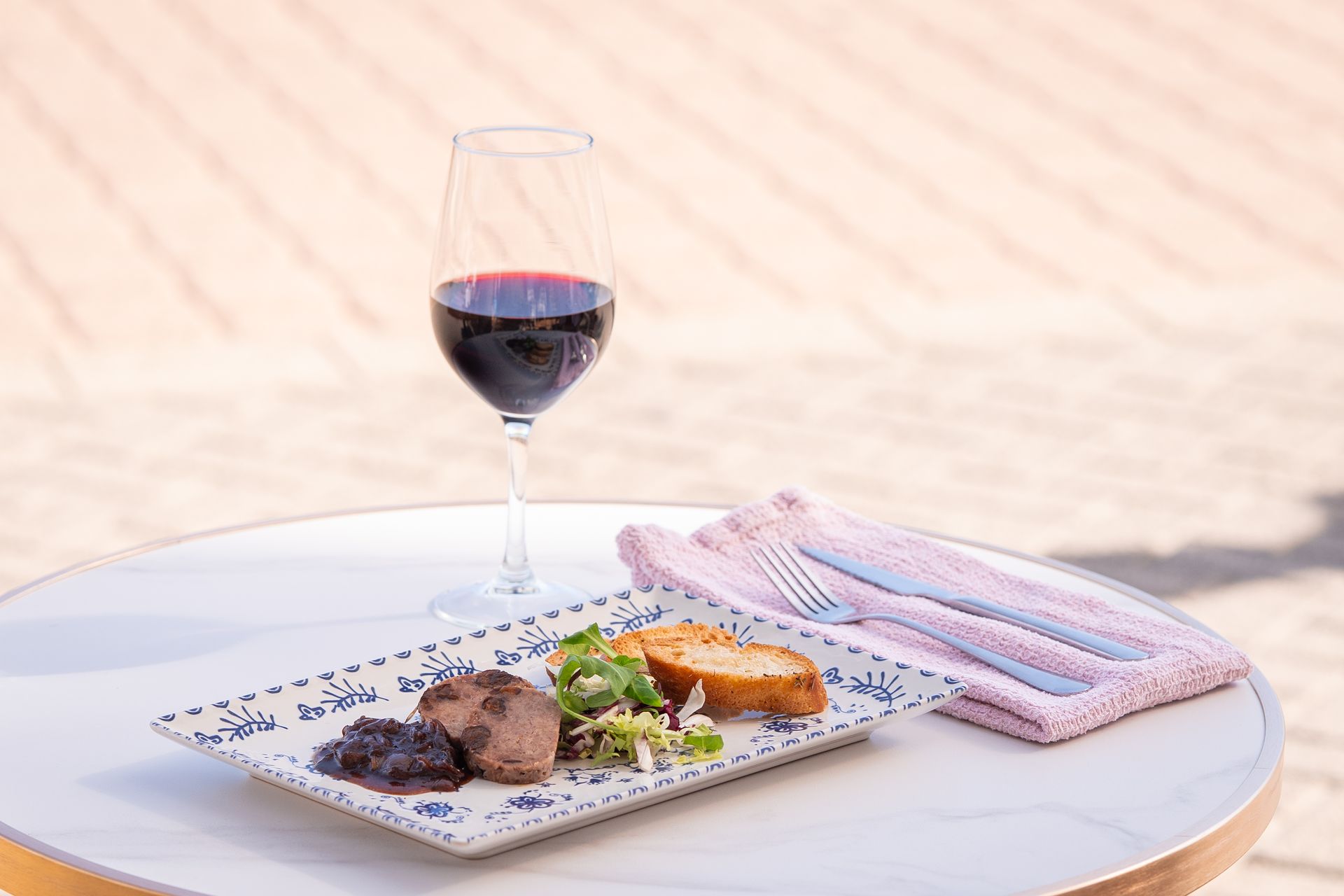 Mesa preparada para una comida: vino, plato principal, servilleta y cubiertos. Ambiente al aire libre con luz cálida.