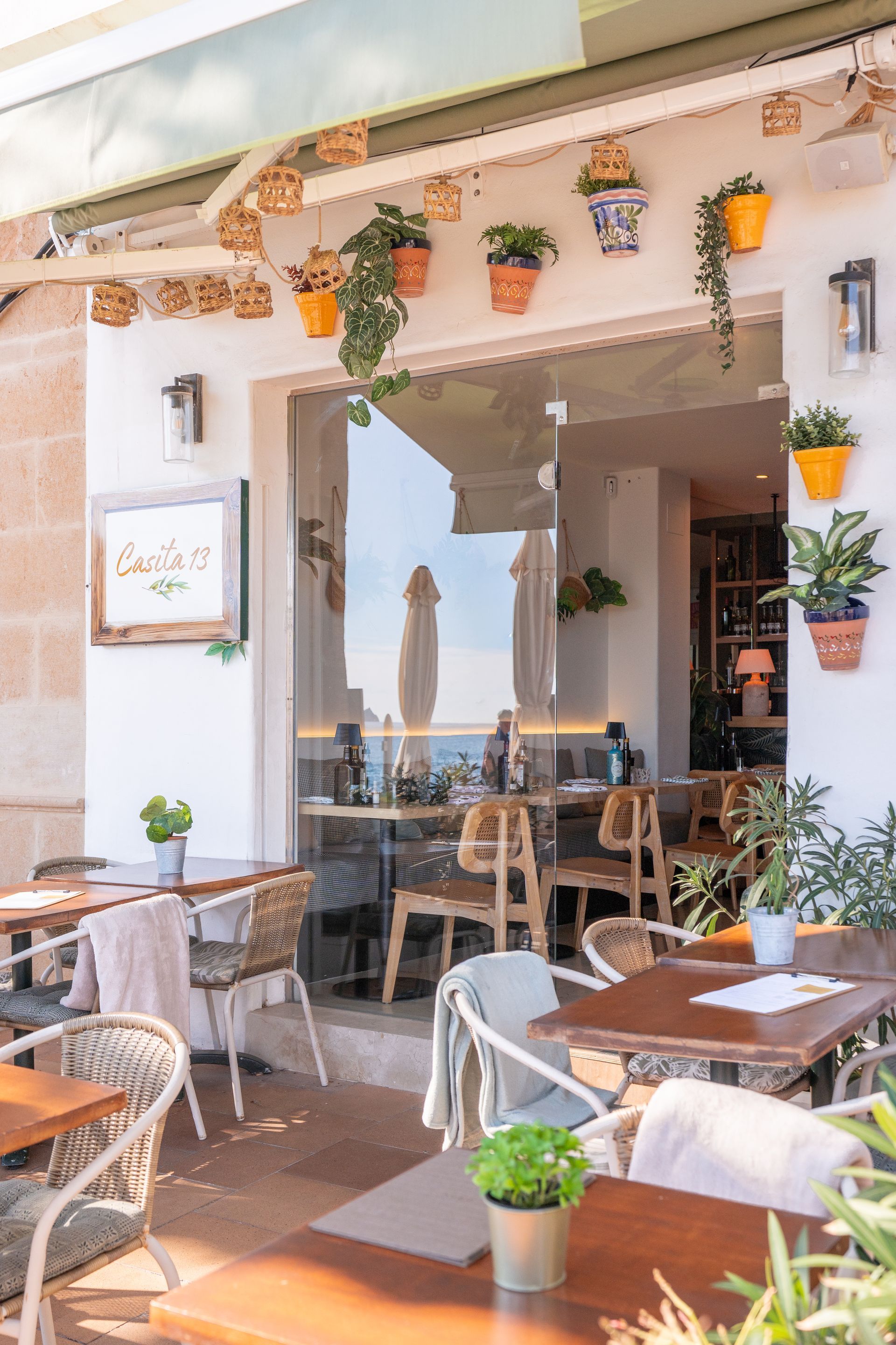 Cafetería al aire libre con mesas y sillas, plantas colgantes y vista al mar.