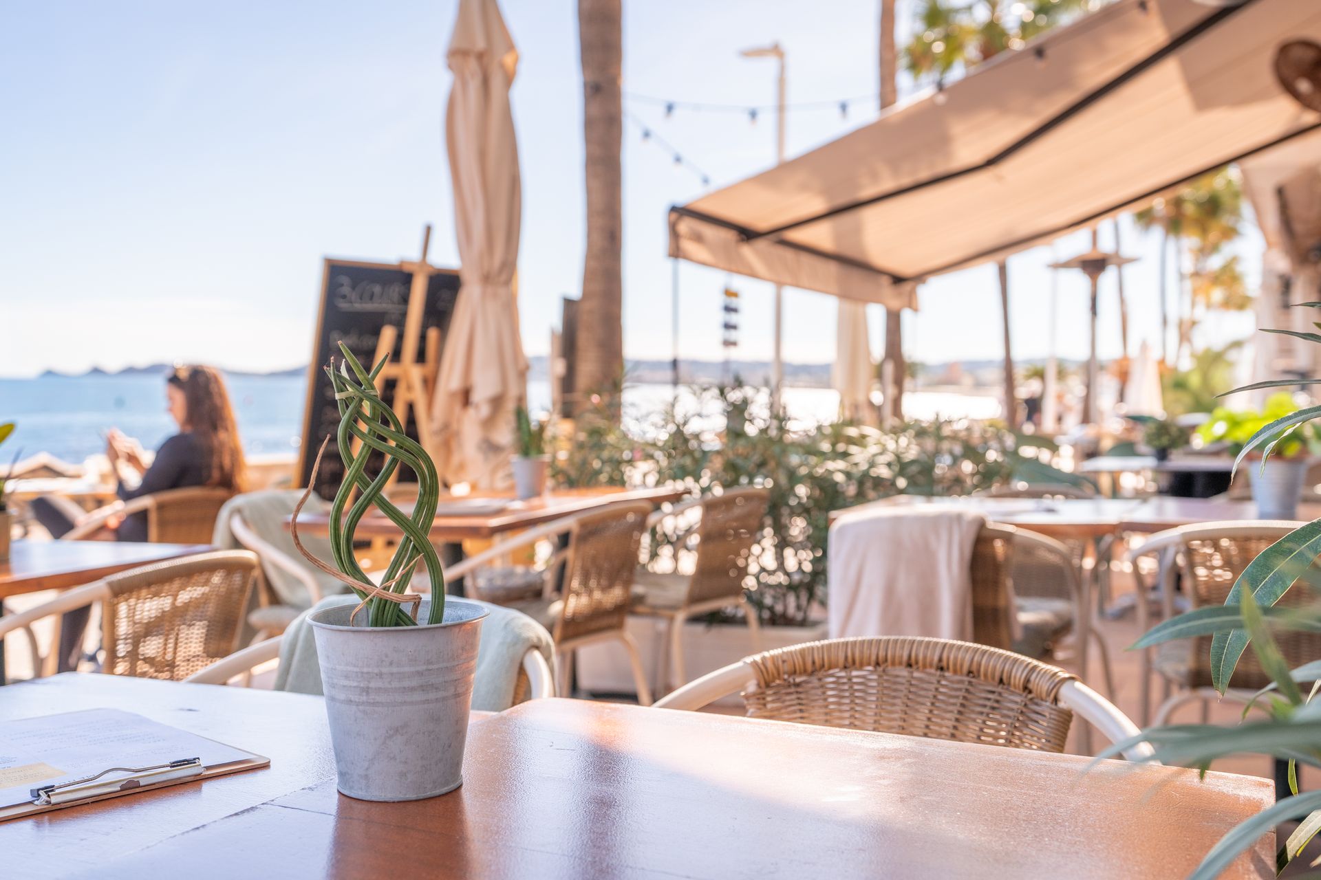 Cafetería al aire libre con mesas, sillas y plantas en maceta, con vista a la playa.