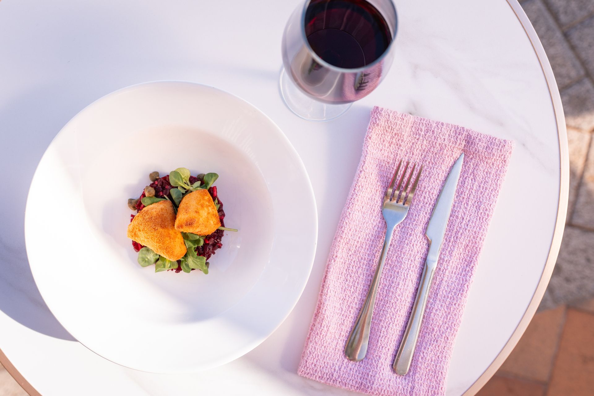 Plato blanco con comida, vino tinto y cubiertos sobre una servilleta rosa sobre una mesa blanca.