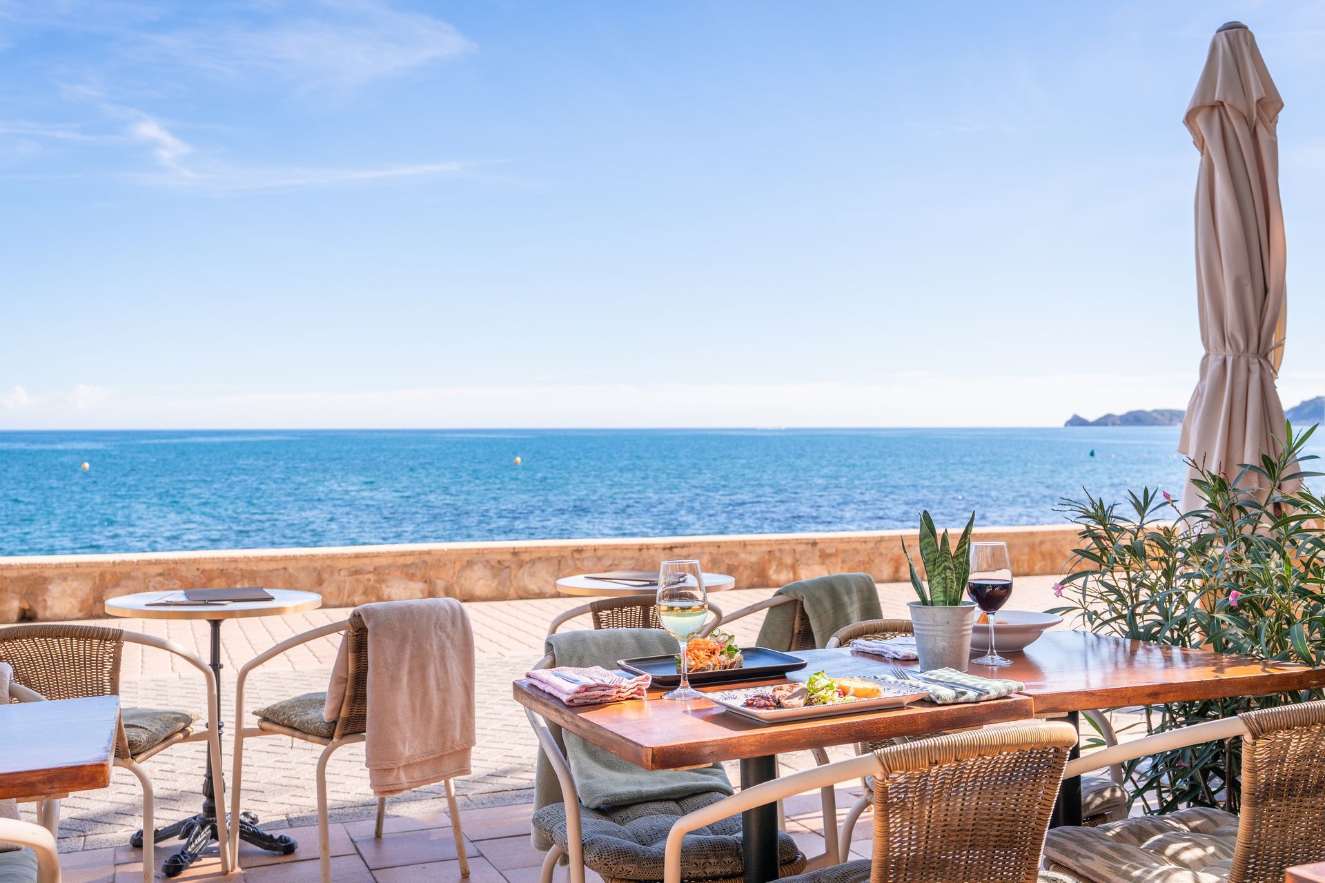 Patio del restaurante con vista al mar con comida, bebidas y una sombrilla cerrada en un día soleado.