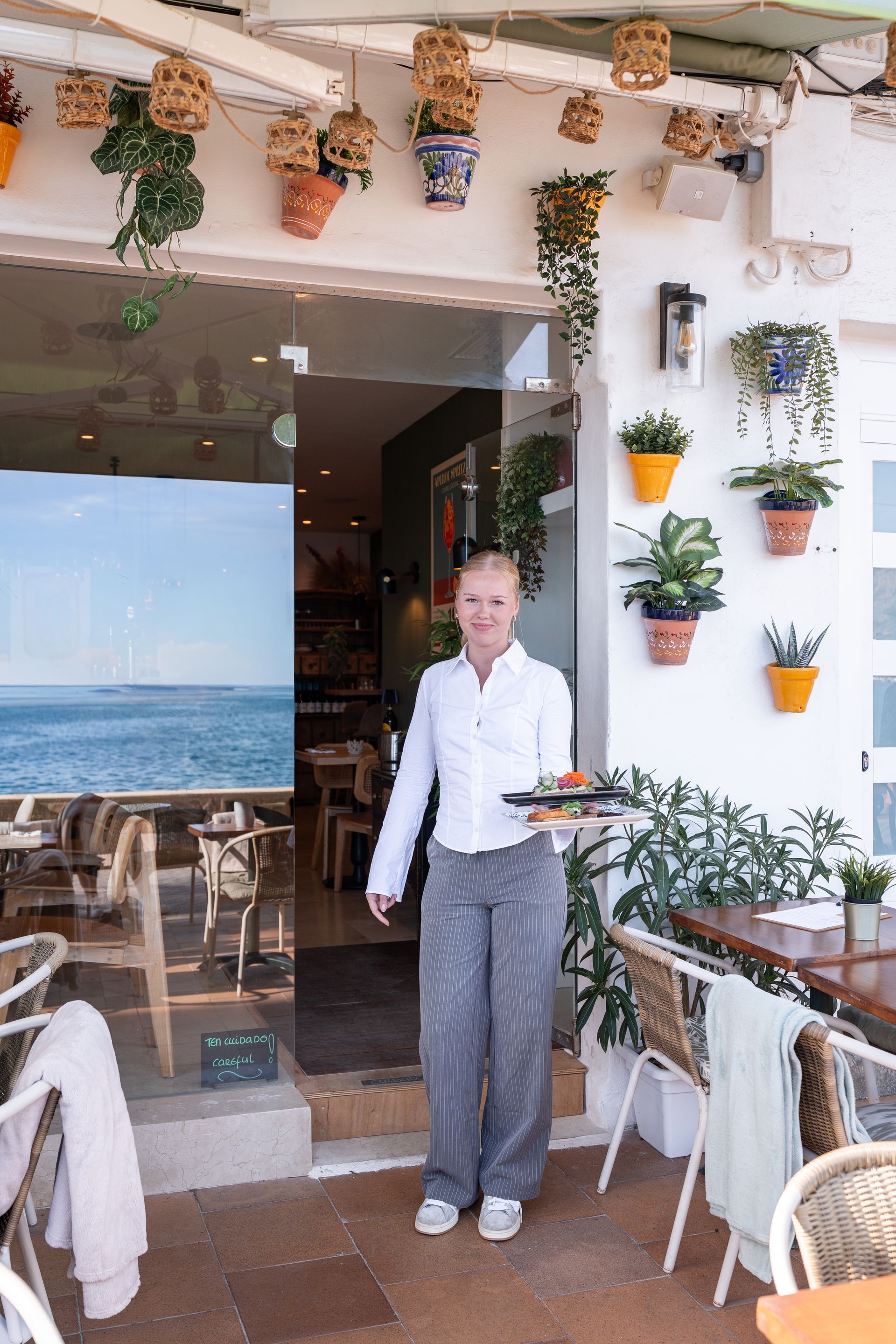 Una mujer con platos sale de un restaurante con terraza. Paredes blancas, plantas y vistas al mar.