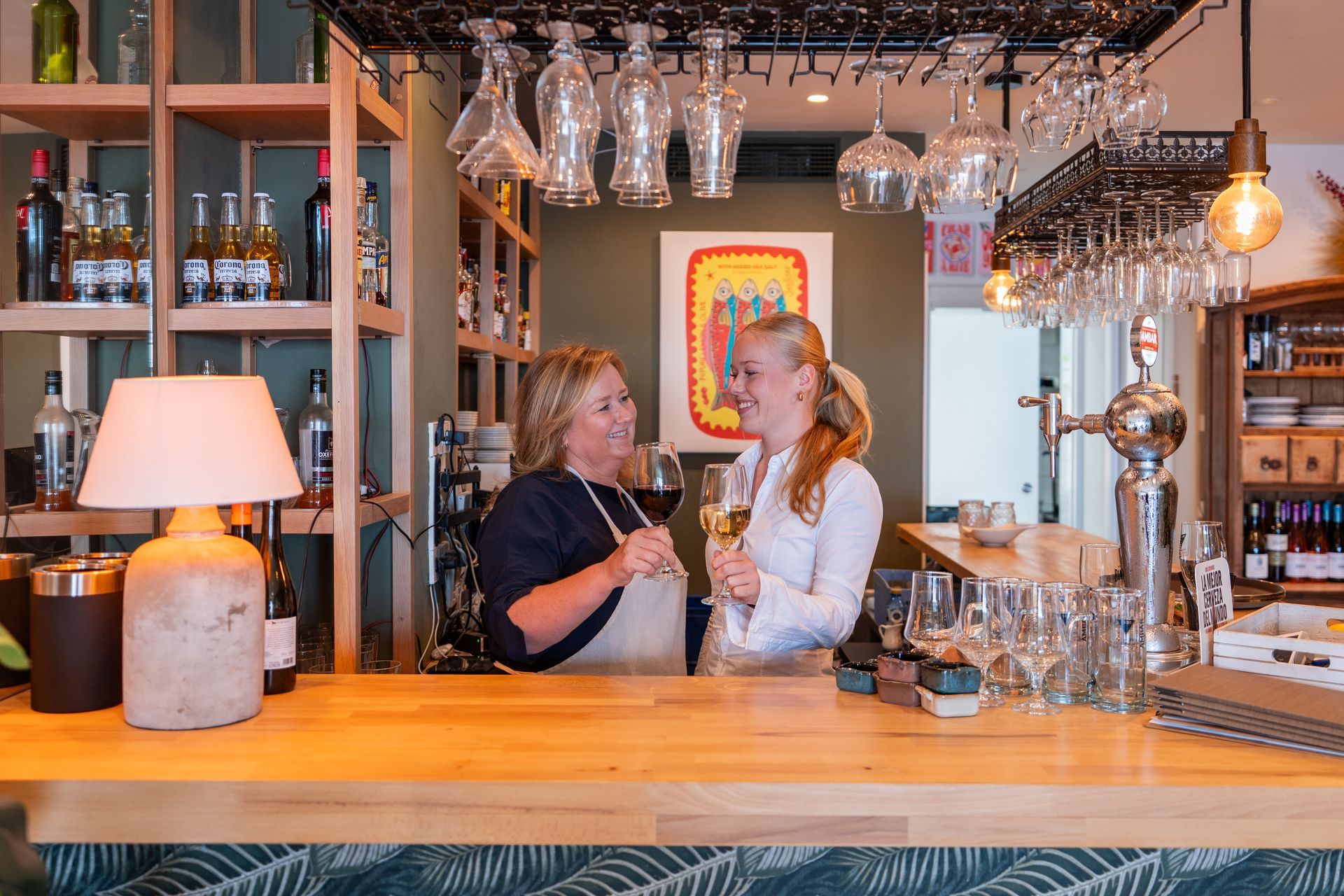 Dos mujeres, una con vino tinto y la otra blanco, en un bar bien iluminado con copas colgando.