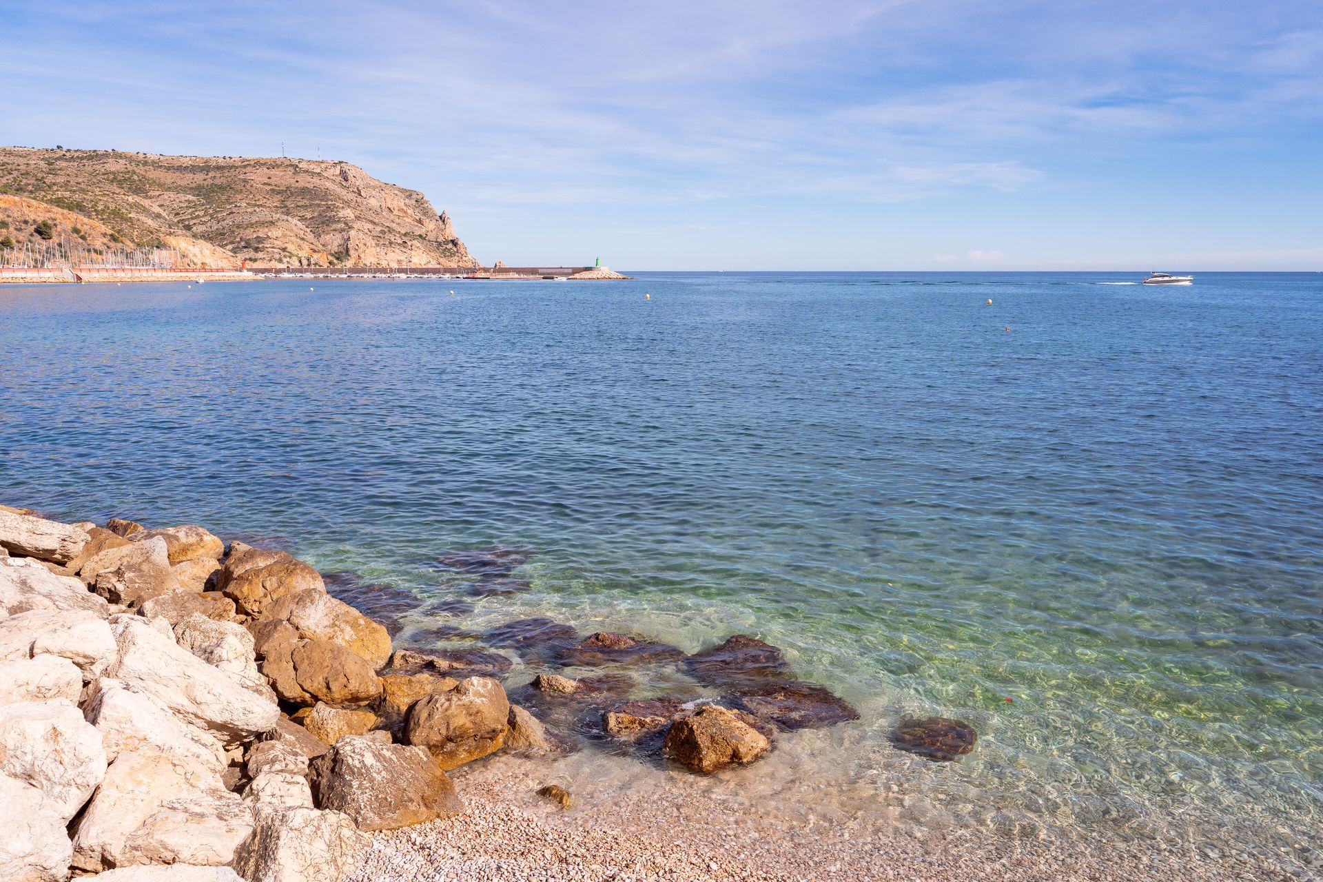 Costa rocosa con agua clara, mar azul y una ladera lejana bajo un cielo brillante.