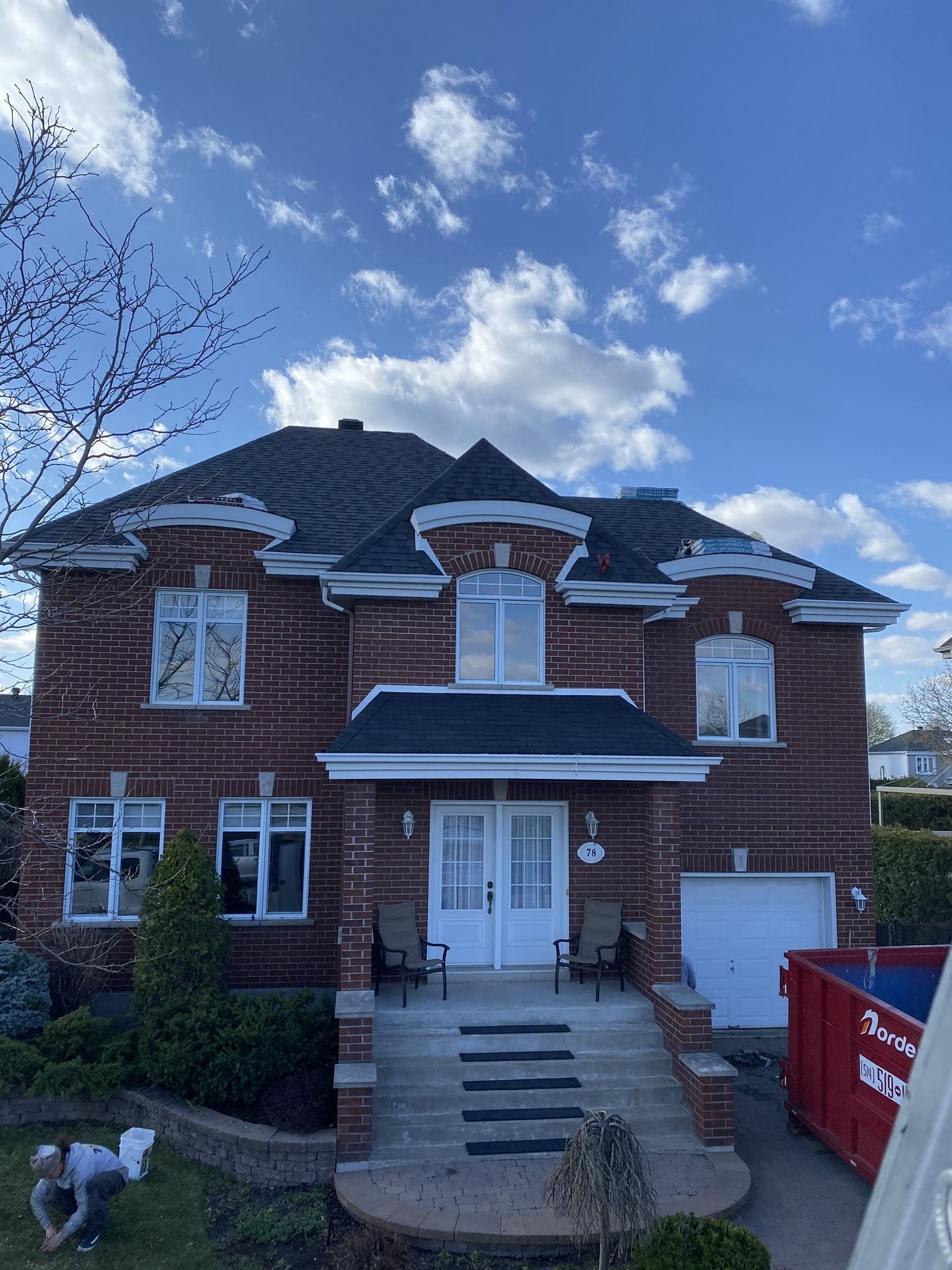Une grande maison en briques avec un toit noir et une benne à ordures rouge devant.