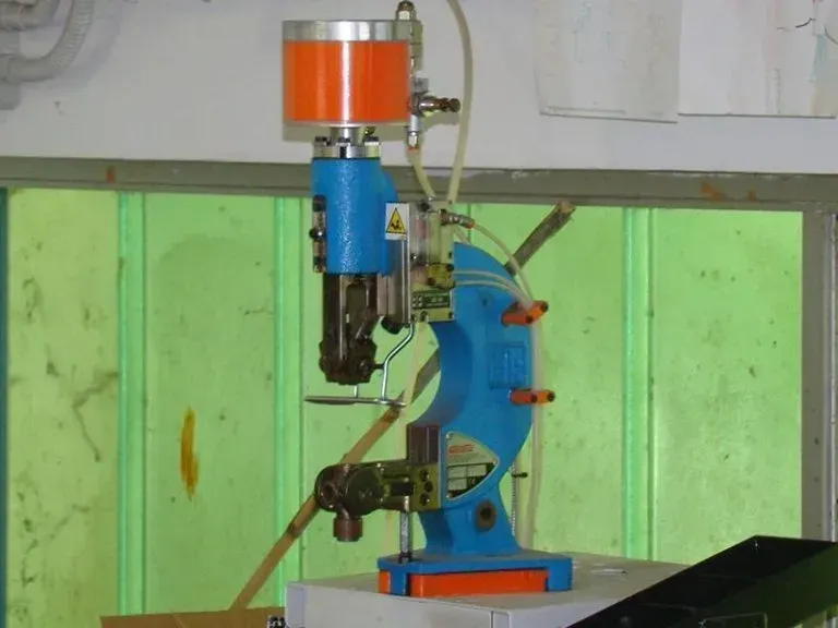 Su un banco da lavoro, contro una parete verde, è posizionata una rivettatrice o una macchina per l'applicazione di occhielli di colore blu e arancione.