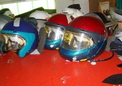 Su una superficie rosso acceso sono appoggiati tre caschi da motociclista, caratterizzati da calotte blu, rosse e argentate con visiere trasparenti.