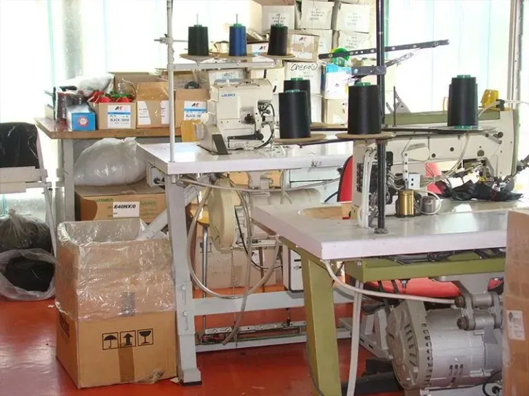 Macchine da cucire industriali con bobine di filo su tavoli da lavoro bianchi in un'officina di fabbrica disordinata.