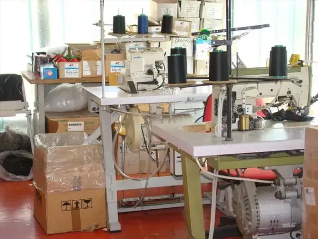 Macchine da cucire industriali con bobine di filo su tavoli da lavoro bianchi in un'officina di fabbrica disordinata.