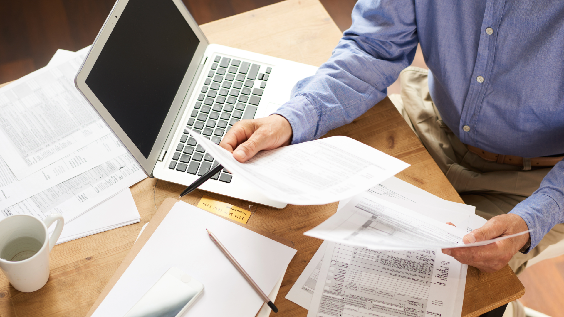 A person in a blue shirt sits at a desk with a laptop, reviewing multiple documents and financial papers.