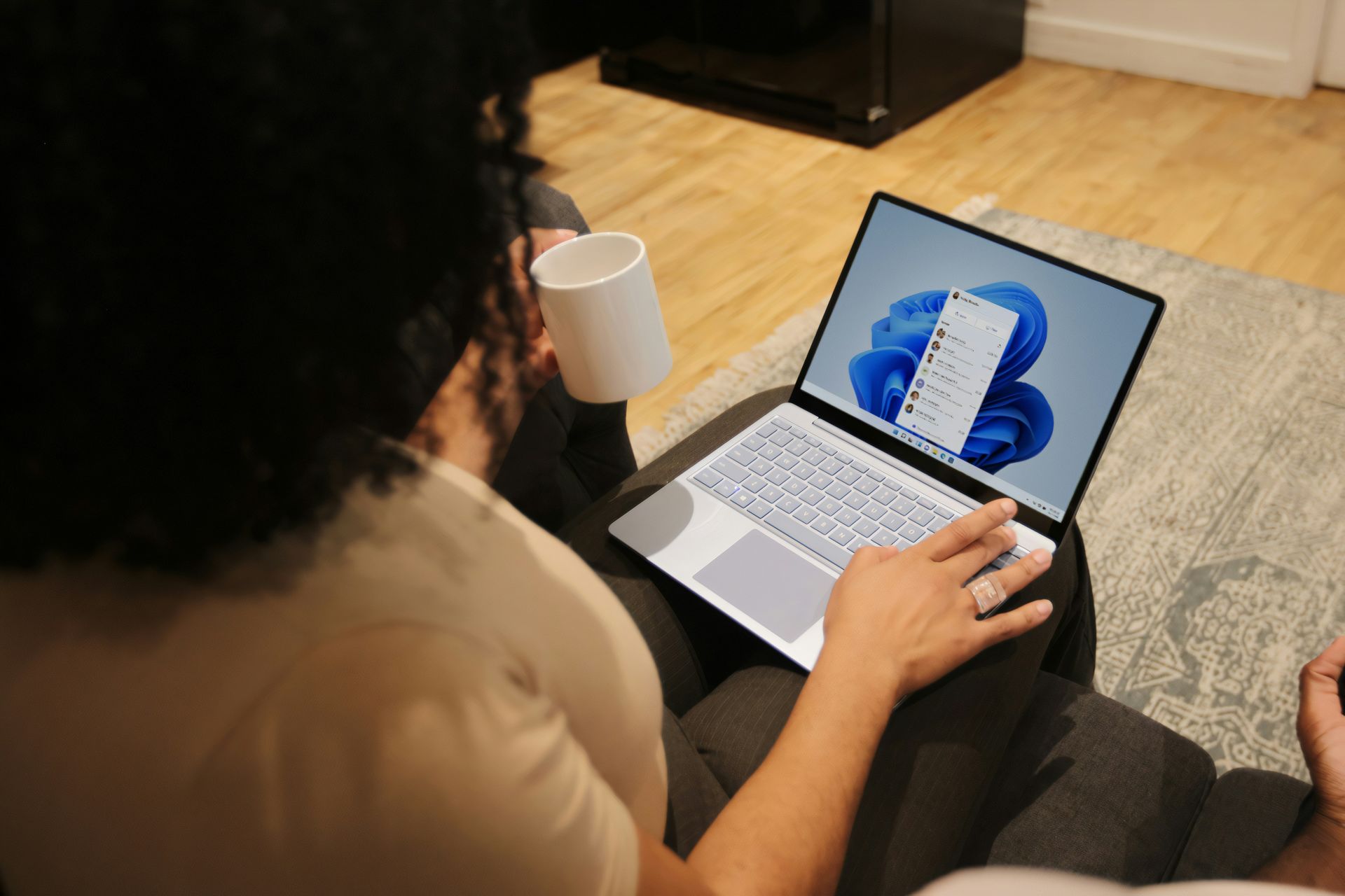 Person using a laptop on a couch, holding a mug. The laptop screen displays a Windows 11 interface.