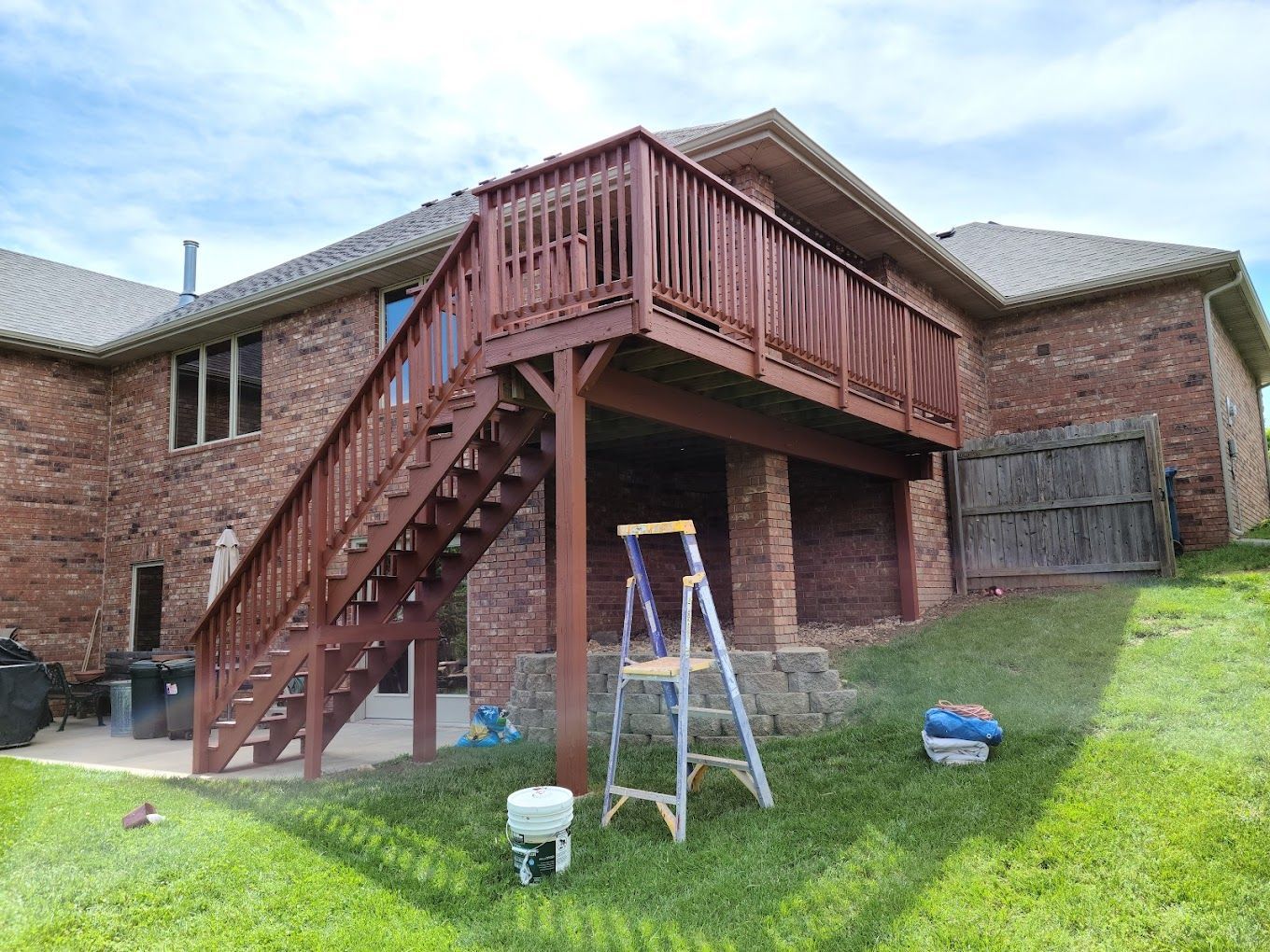 A wooden deck with stairs and a ladder in front of a brick house.