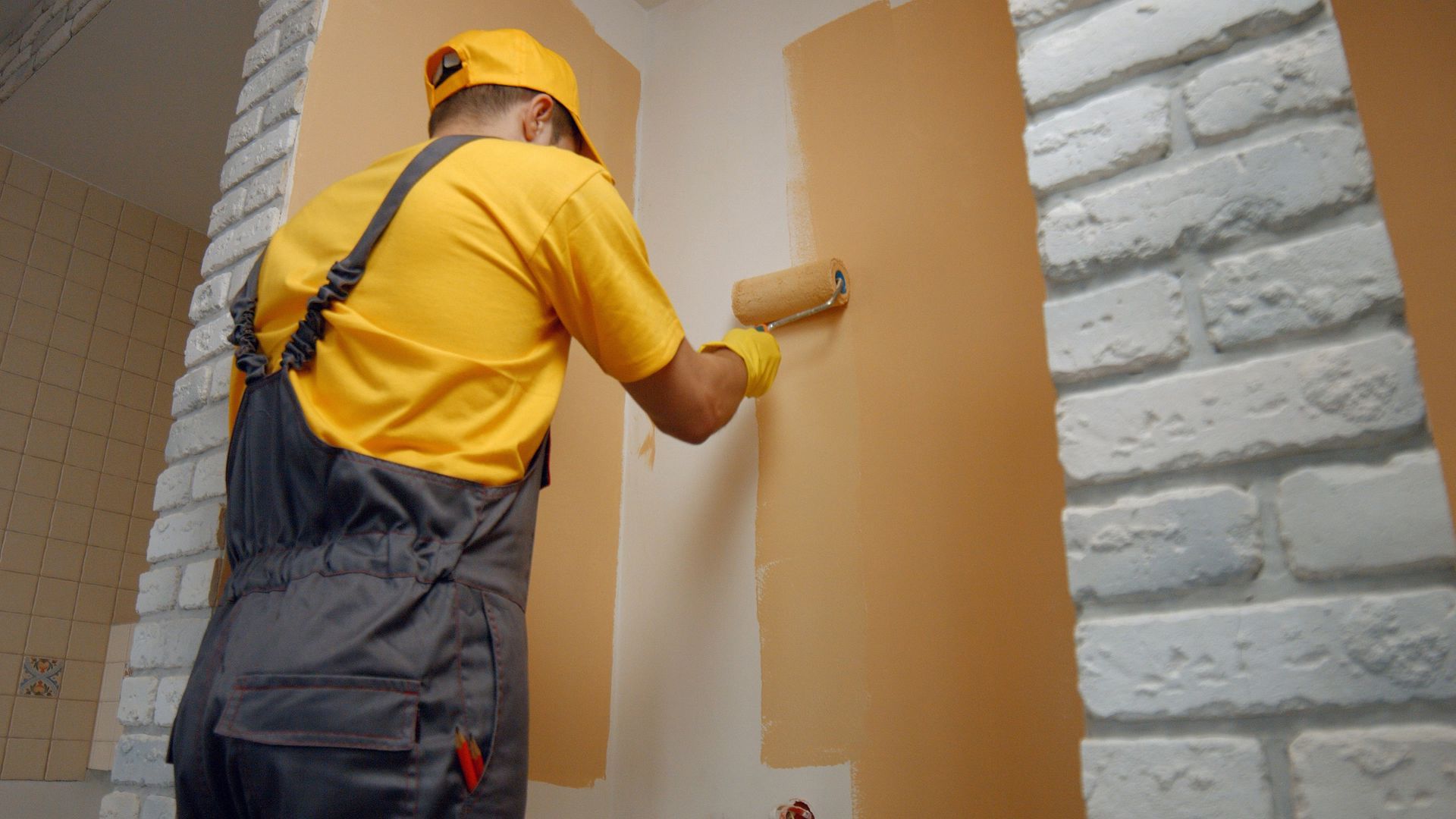 Painter in yellow overalls applying white paint to a wall with a roller, beside a white brick surface