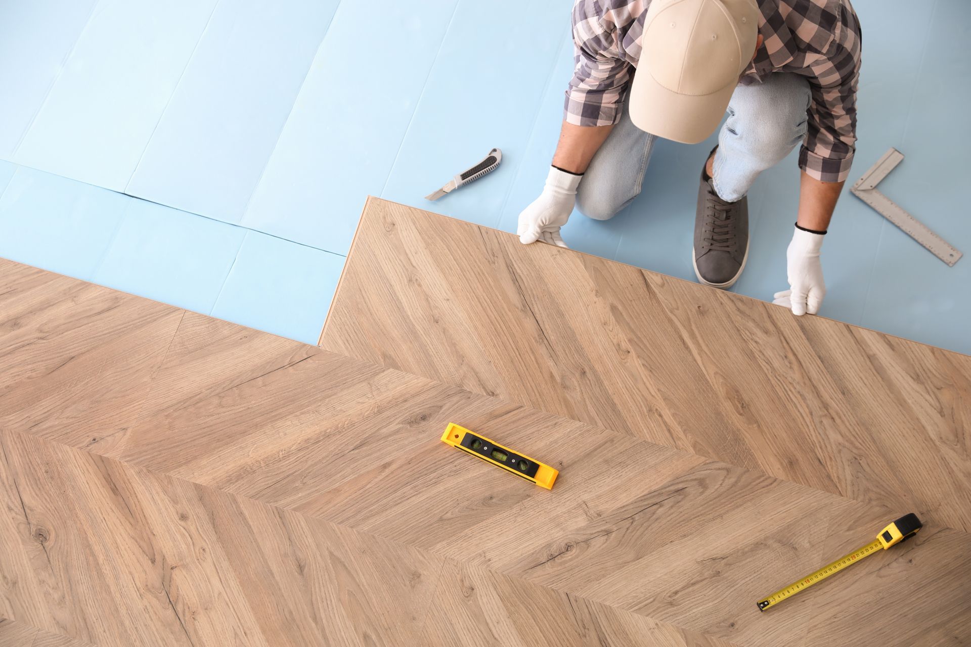 Person laying herringbone wood floor beside a blue underlayment, with measuring tools nearby.