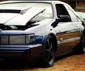 A blue car with the hood up is parked in the grass