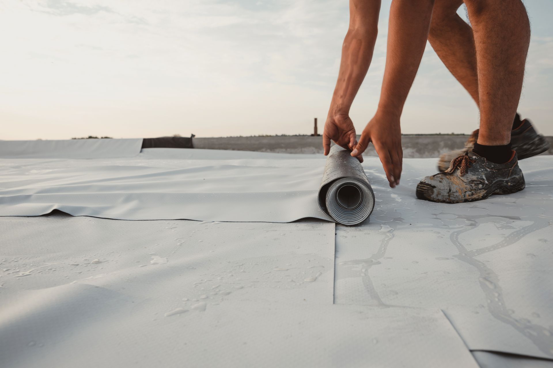 Person rolling out white roofing material on a flat roof, hands visible, clear sky.
