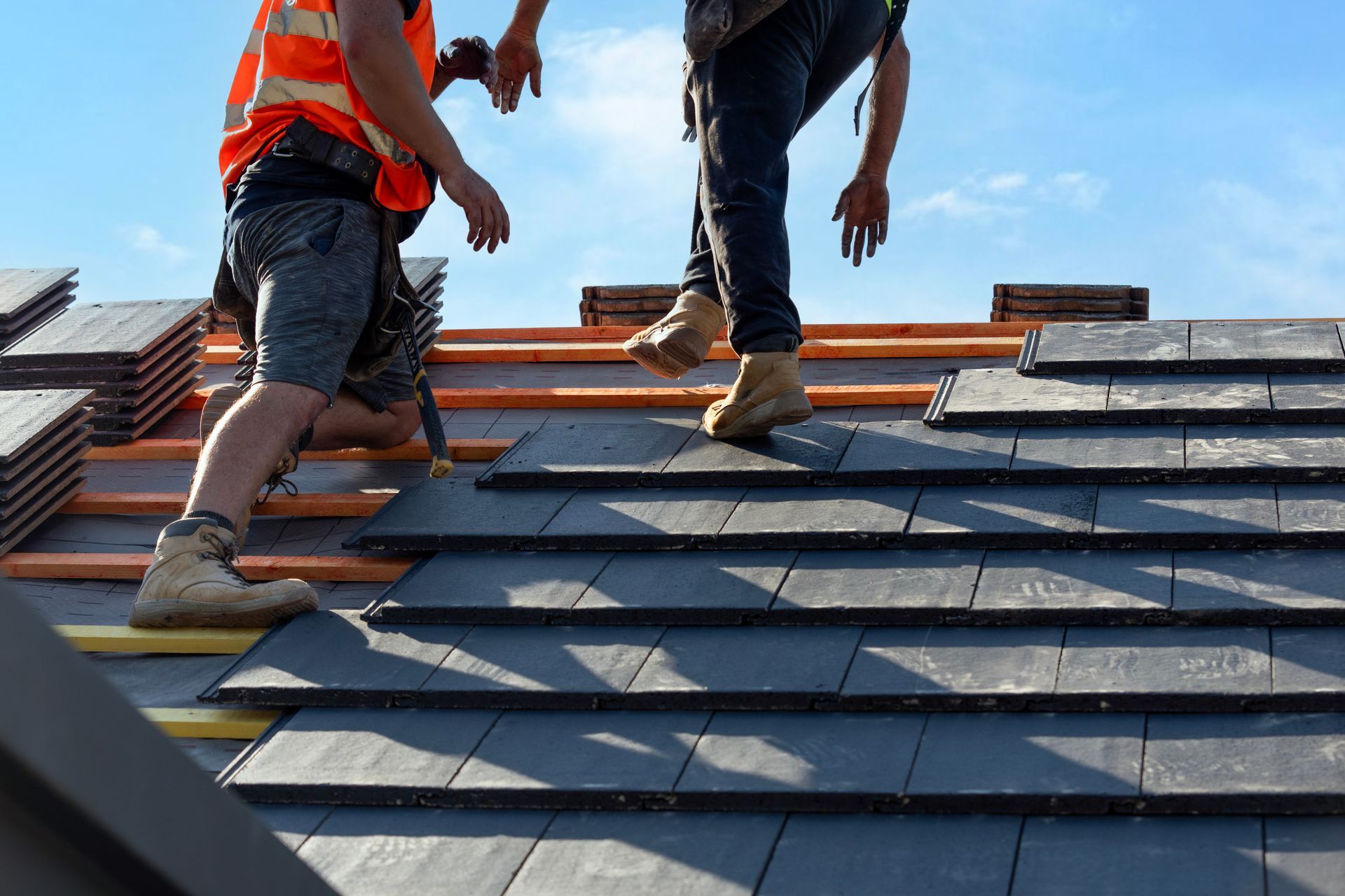 Two workers installing dark gray roof tiles on a sunny day.