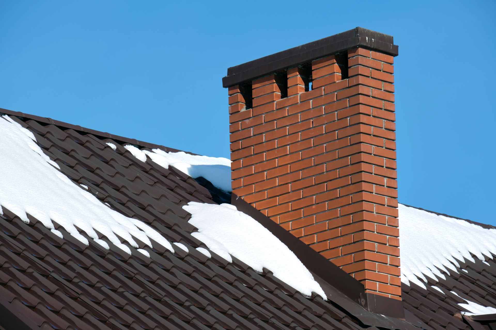 Brick chimney on a brown roof covered with snow, against a blue sky.