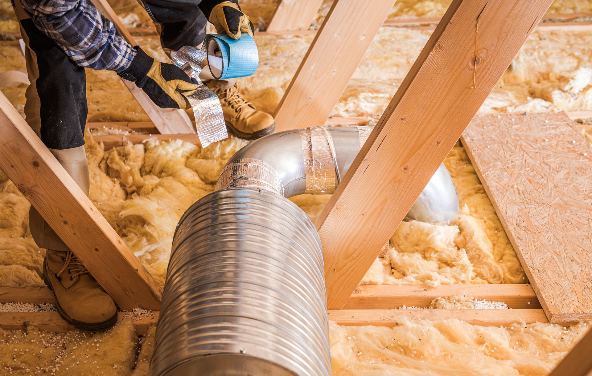 Person installing ductwork in an attic, surrounded by insulation and wooden beams.