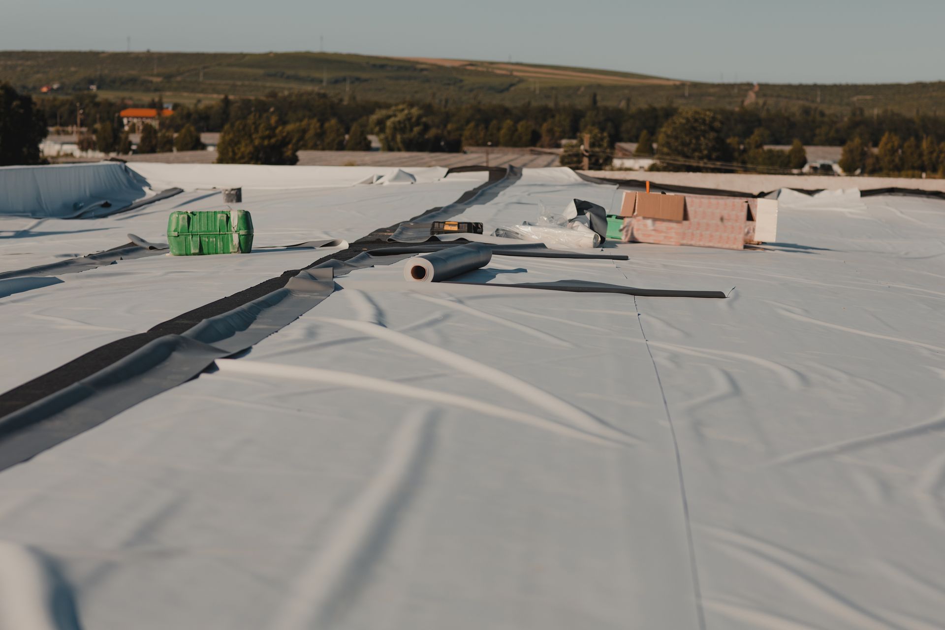 White roofing material being installed on a flat roof, with equipment and a backdrop of trees and a hill.