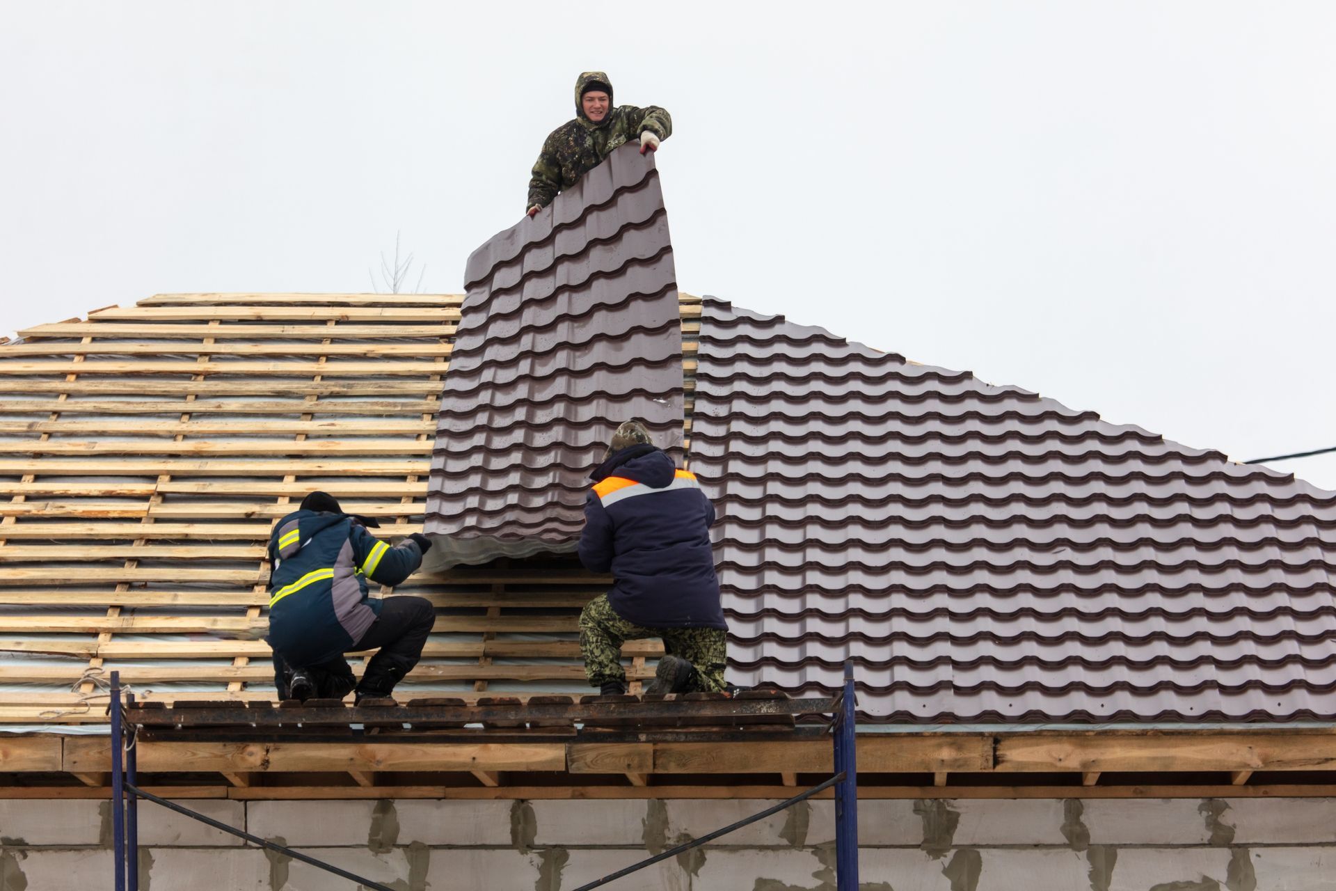 Three construction workers installing a metal roof on a house, working on a cloudy day.