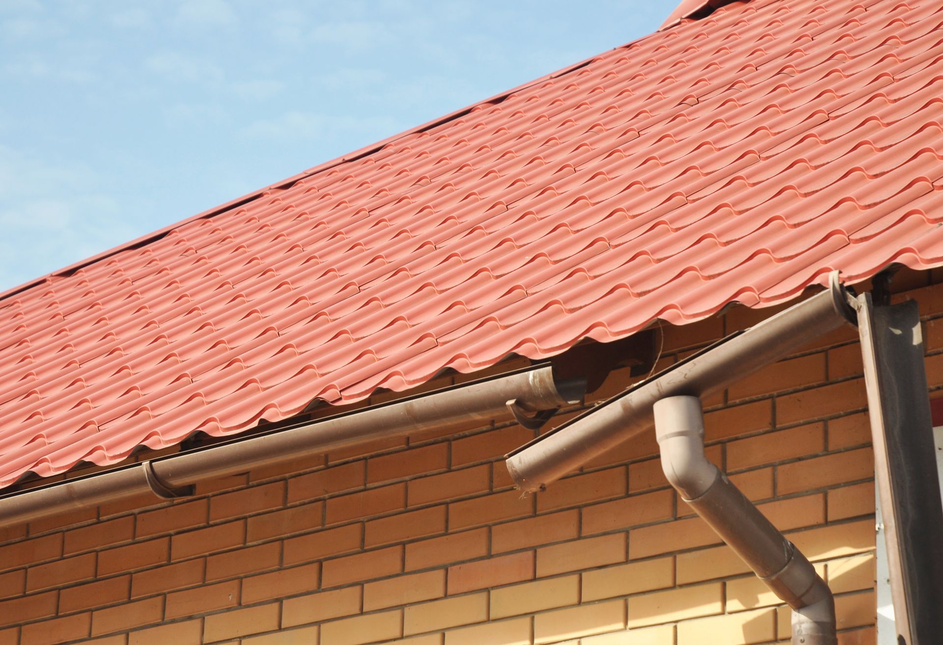 Red corrugated metal roof with brown gutters and downspout on a brick building.
