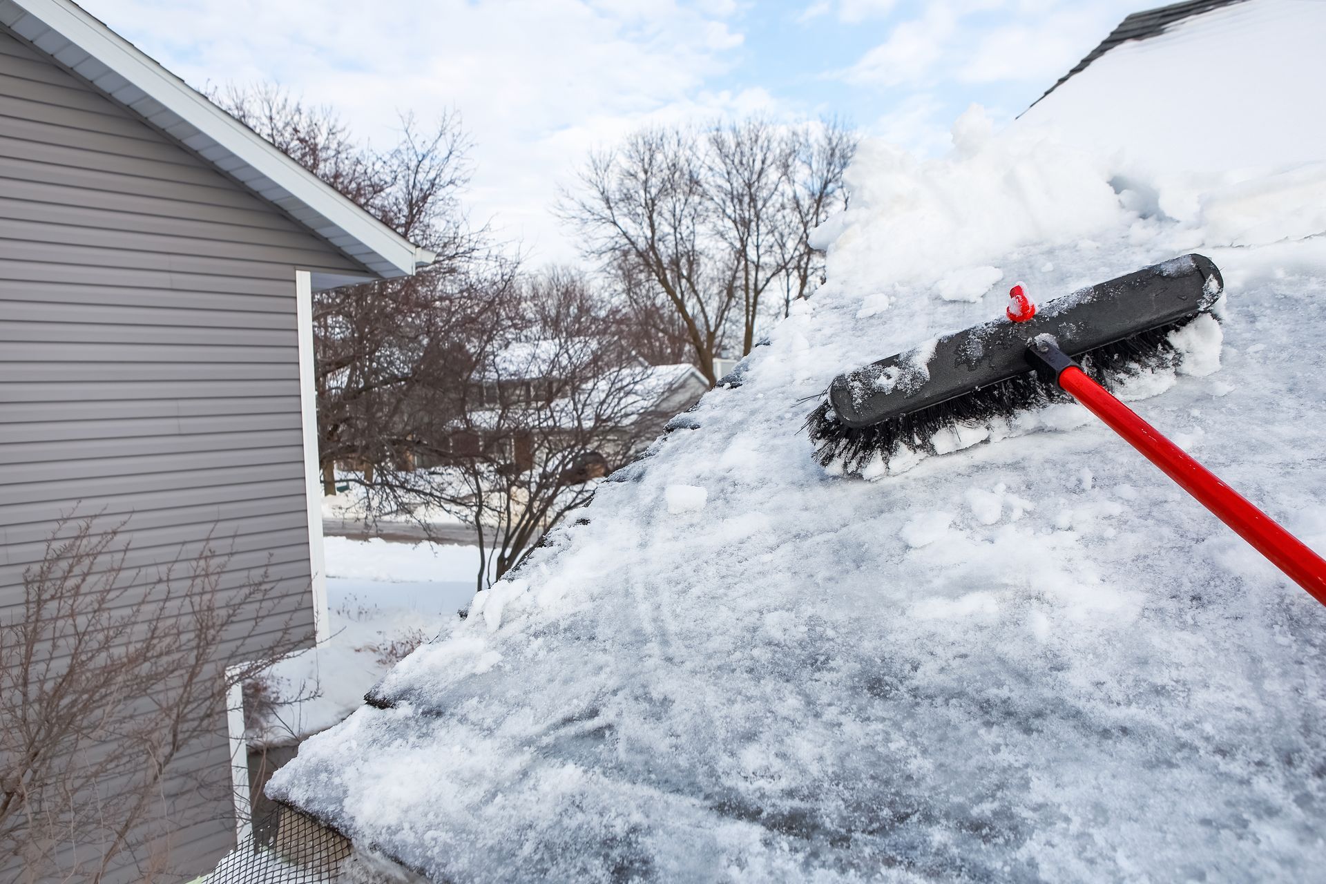 A person using a red-handled snow roof rake to remove snow from a house roof on a cloudy day.