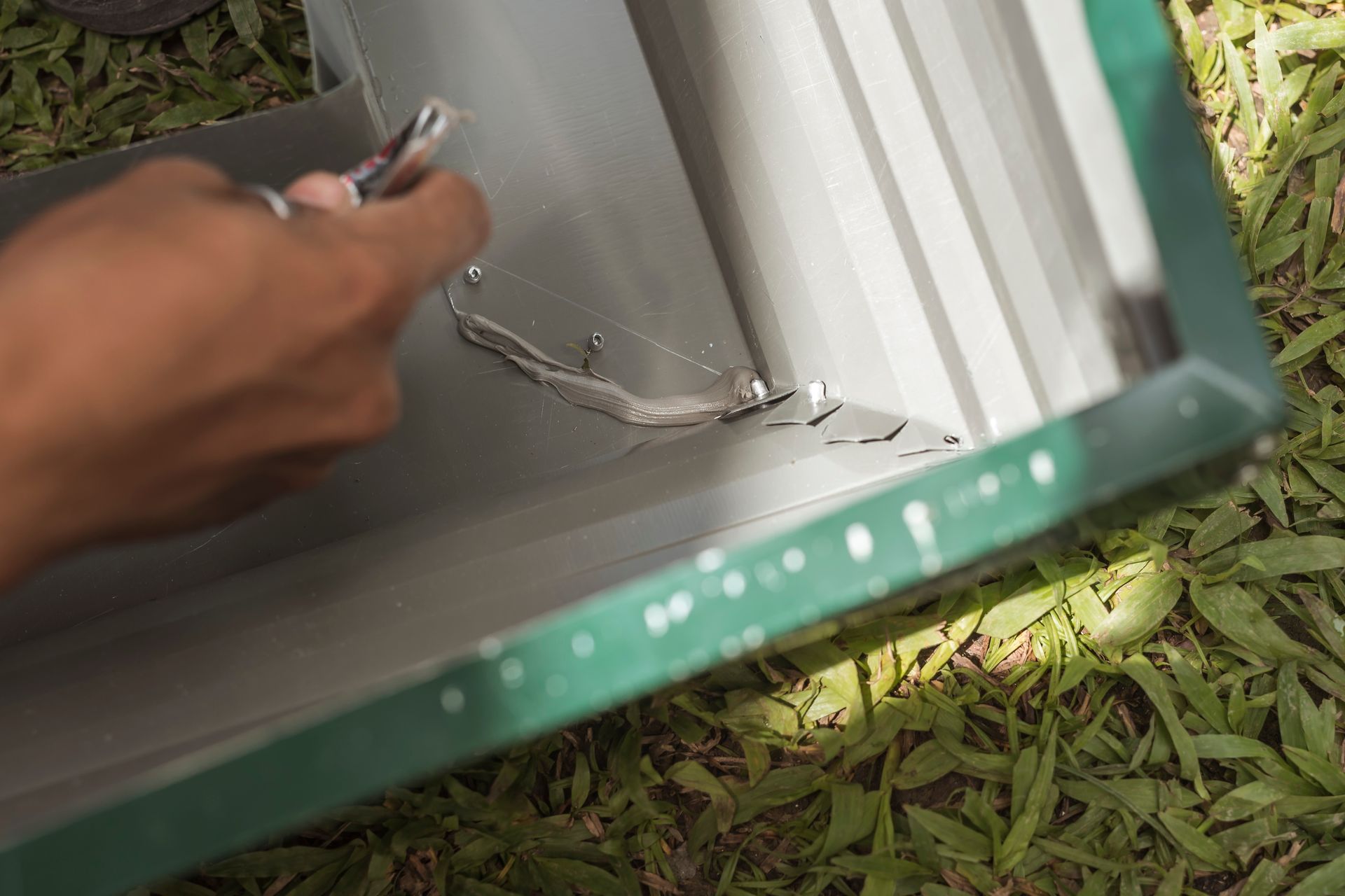 Hand using pliers to fix a metallic, green-framed container with gray interior, outdoors on grass.