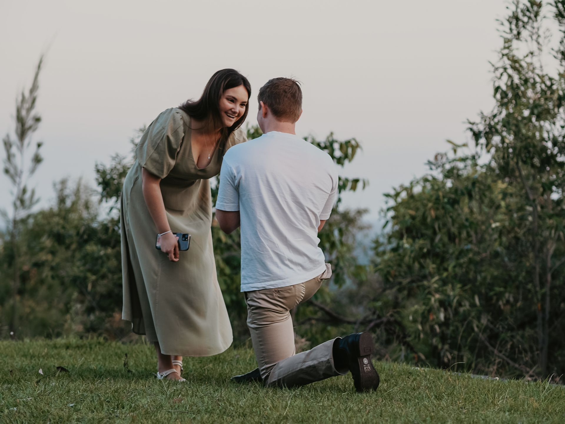 Man kneeling proposing to woman; she smiles, holding a small box. Outdoors on a grassy hill, trees in the background.