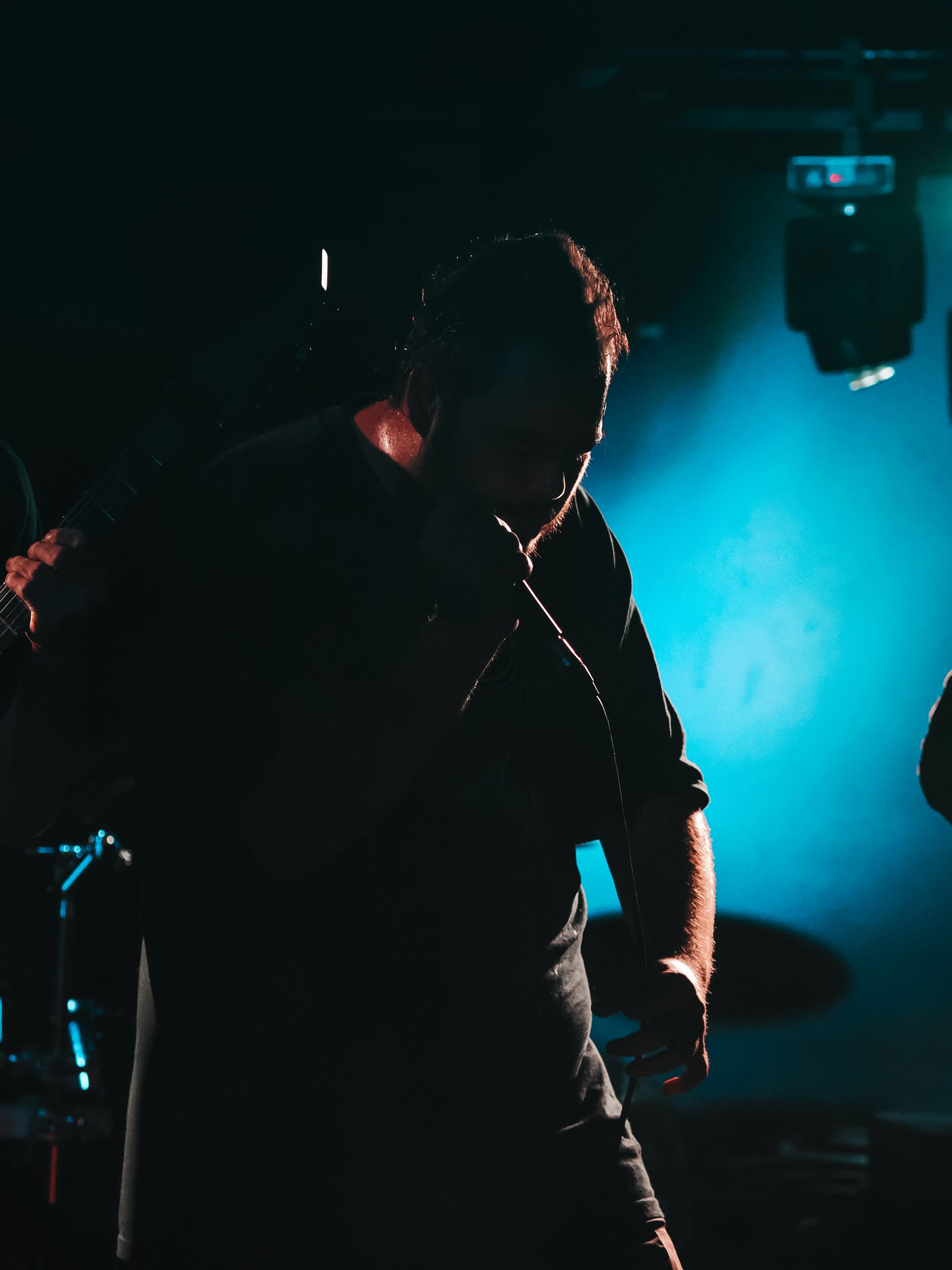 Man singing on stage, backlit with blue light. He holds a microphone, silhouetted with dark clothing.