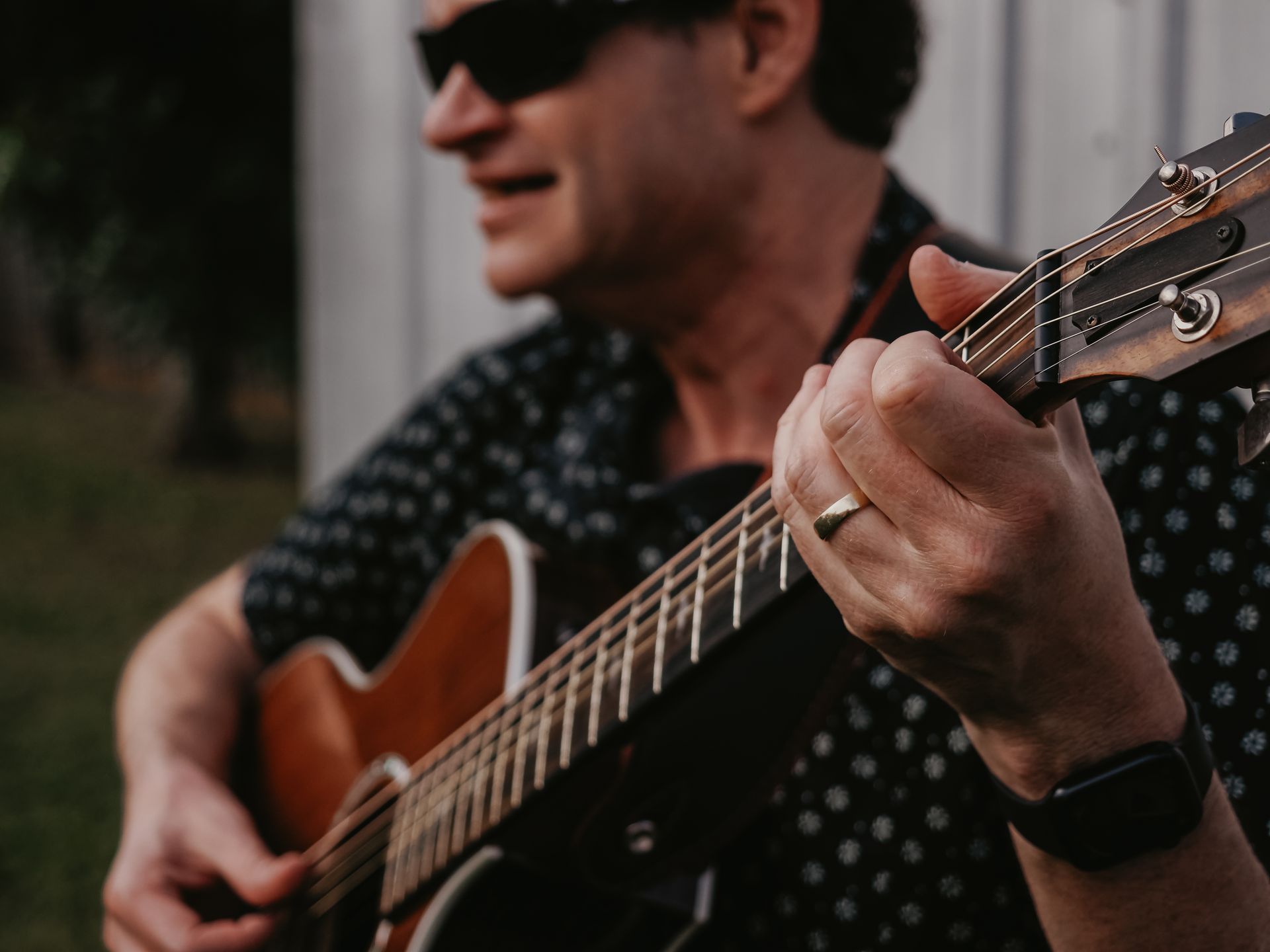 Man with sunglasses playing an acoustic guitar outdoors. His hand strums the strings, a wedding ring is on his finger.