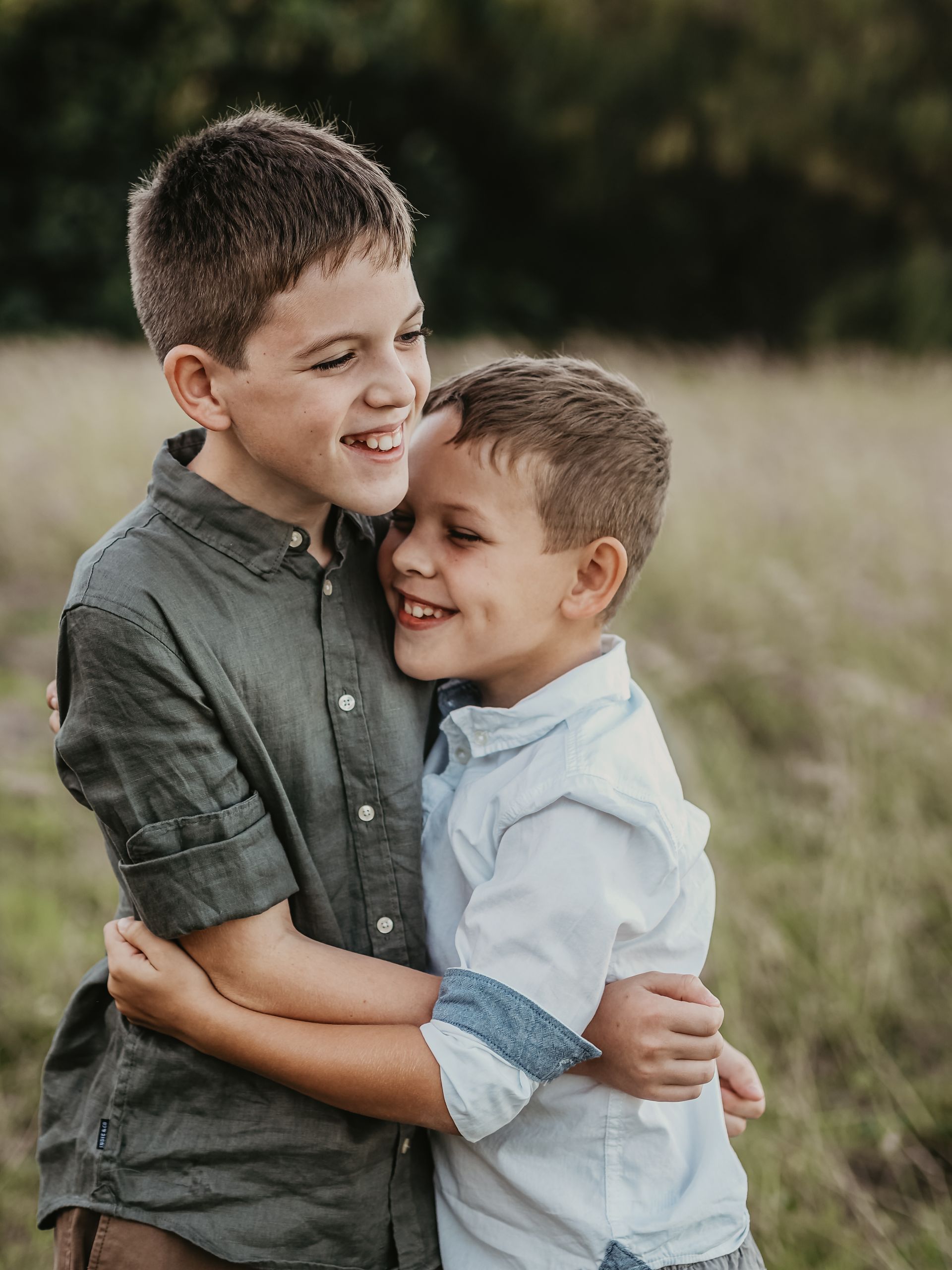 Two young boys hugging and smiling in a field, one wearing a green shirt and the other a blue shirt.