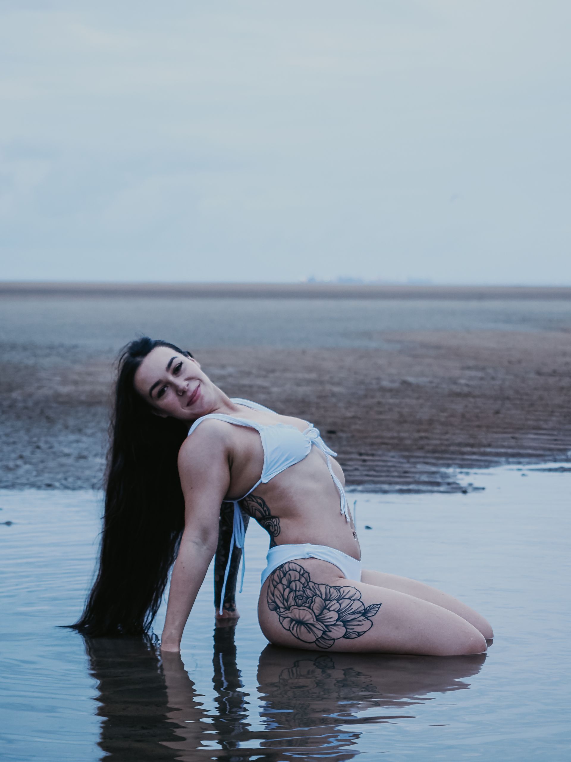 Woman with long dark hair and a bikini kneels in shallow water on a beach, looking at the camera and smiling.