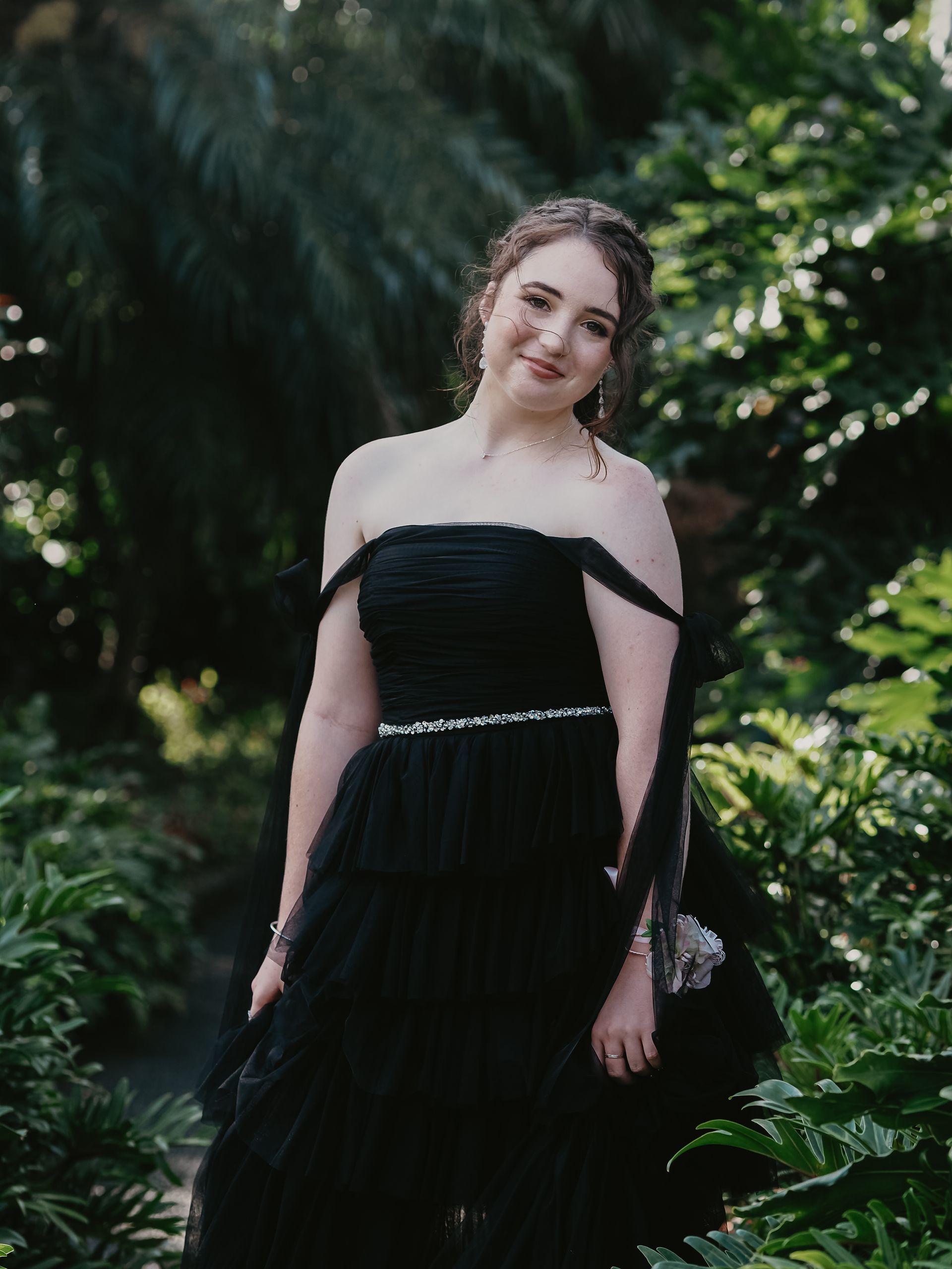 A young woman in a black off-the-shoulder gown smiles, posing in a lush, green garden.